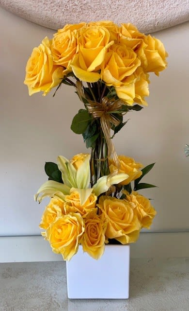 Tall arrangement of yellow roses and pale lilies in a white square vase
