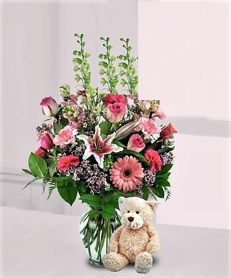 Pink mixed flower arrangement with lilies, roses, and gerbera daisies in a glass vase with a tan teddy bear.