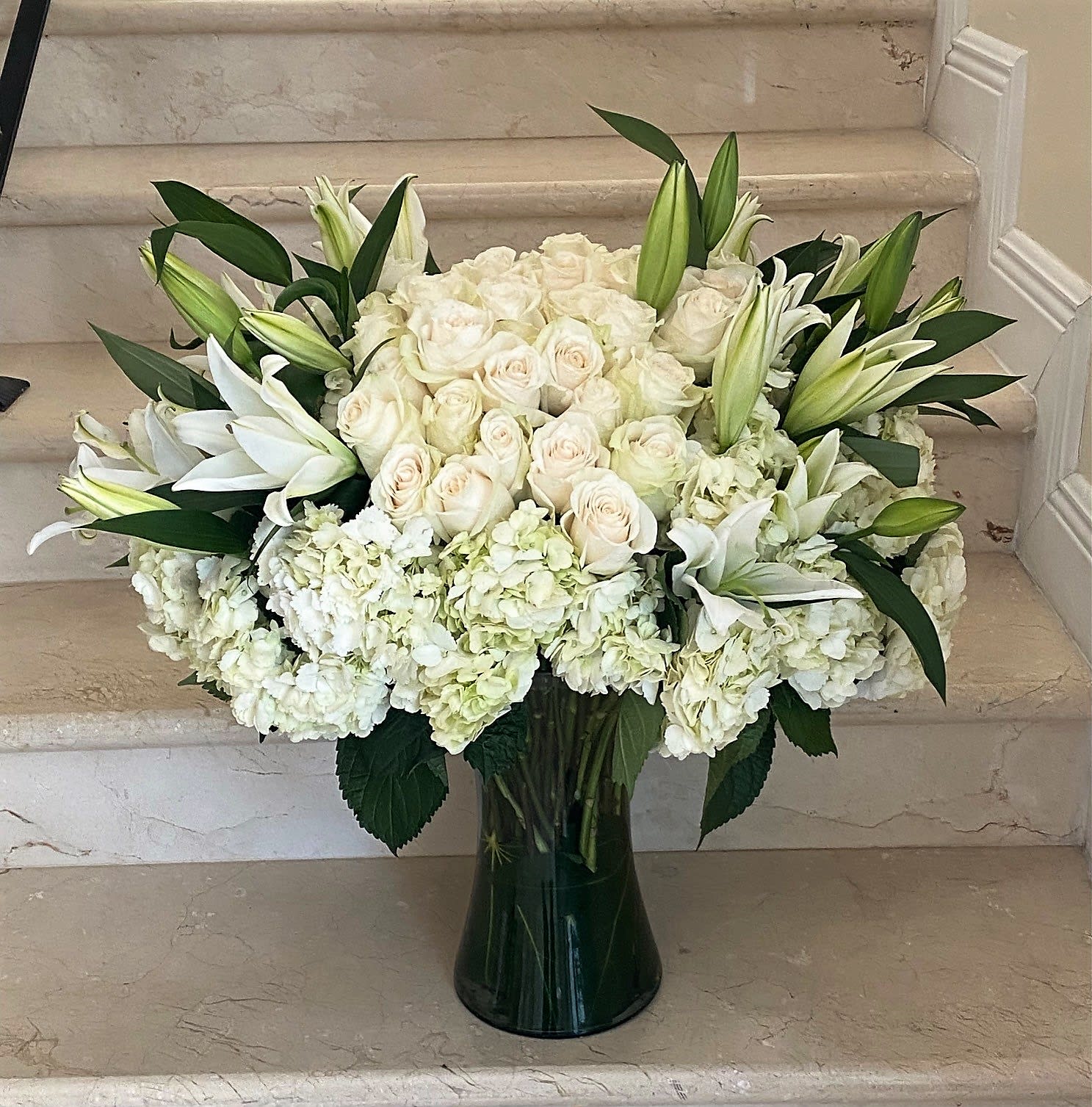 Large white arrangement of roses, lilies, and hydrangeas in a tall clear glass vase