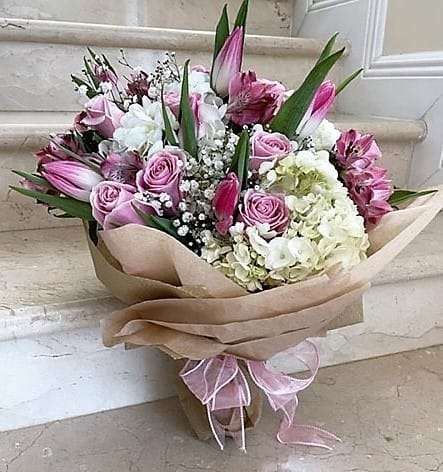 Hand-tied bouquet of pink roses, white hydrangeas, and pink lilies wrapped in kraft paper with a pink ribbon