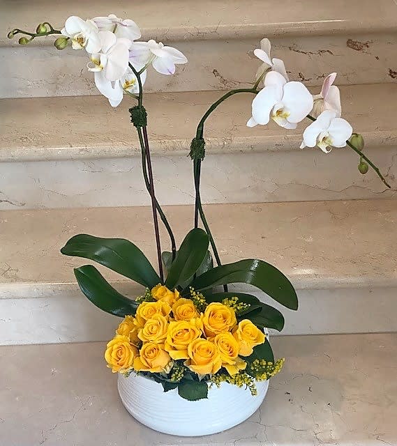White orchids and yellow roses arranged in a white ceramic bowl with lush green leaves