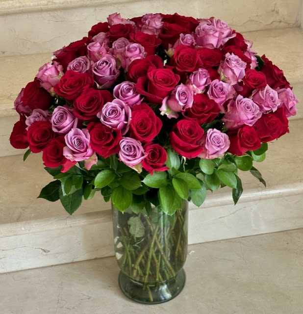 Large arrangement of red and lavender roses in a clear glass vase with a lush green collar