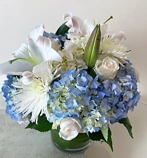 Round arrangement of blue hydrangeas, white lilies, roses, and mums in a clear glass vase