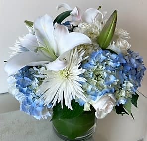 Compact arrangement of white lilies, mums, and roses with blue hydrangeas in a clear glass vase