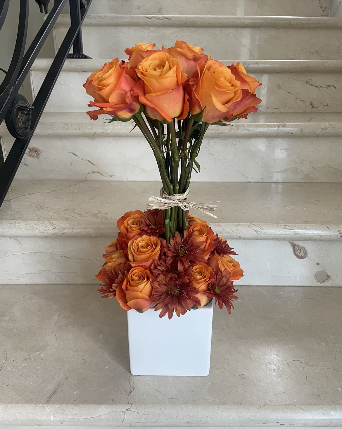 Orange roses and burgundy daisies arranged in a white square vase