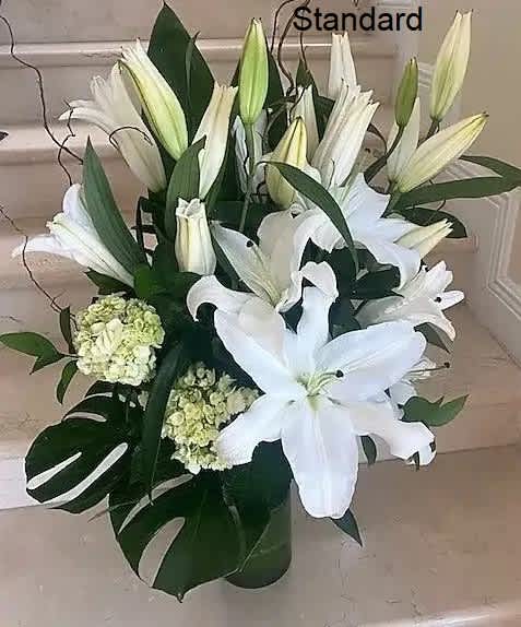 Tall arrangement of white lilies and hydrangeas with monstera leaves in a clear glass vase