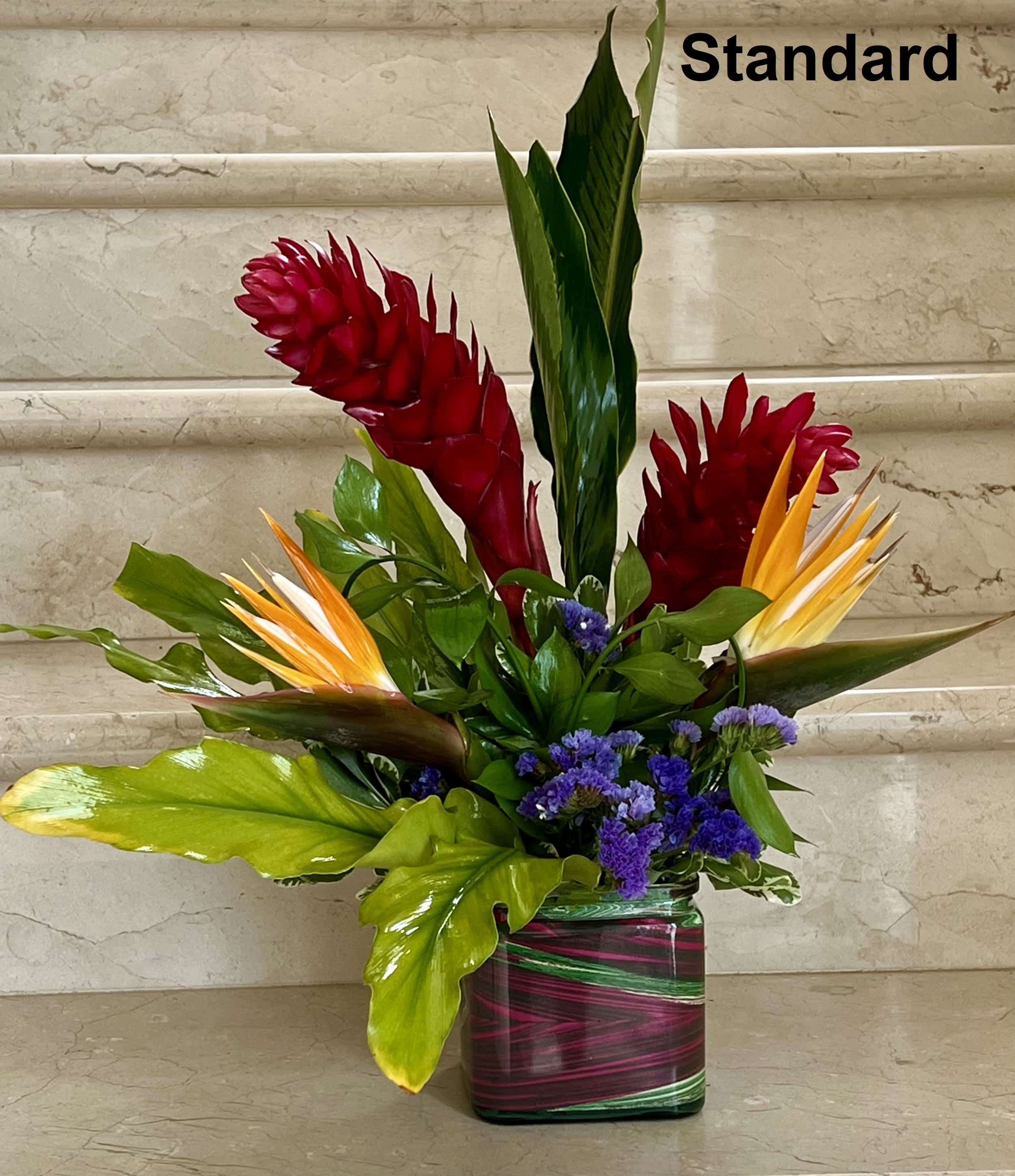 Tall tropical arrangement with red ginger, orange blooms, and purple flowers in a striped square glass vase.