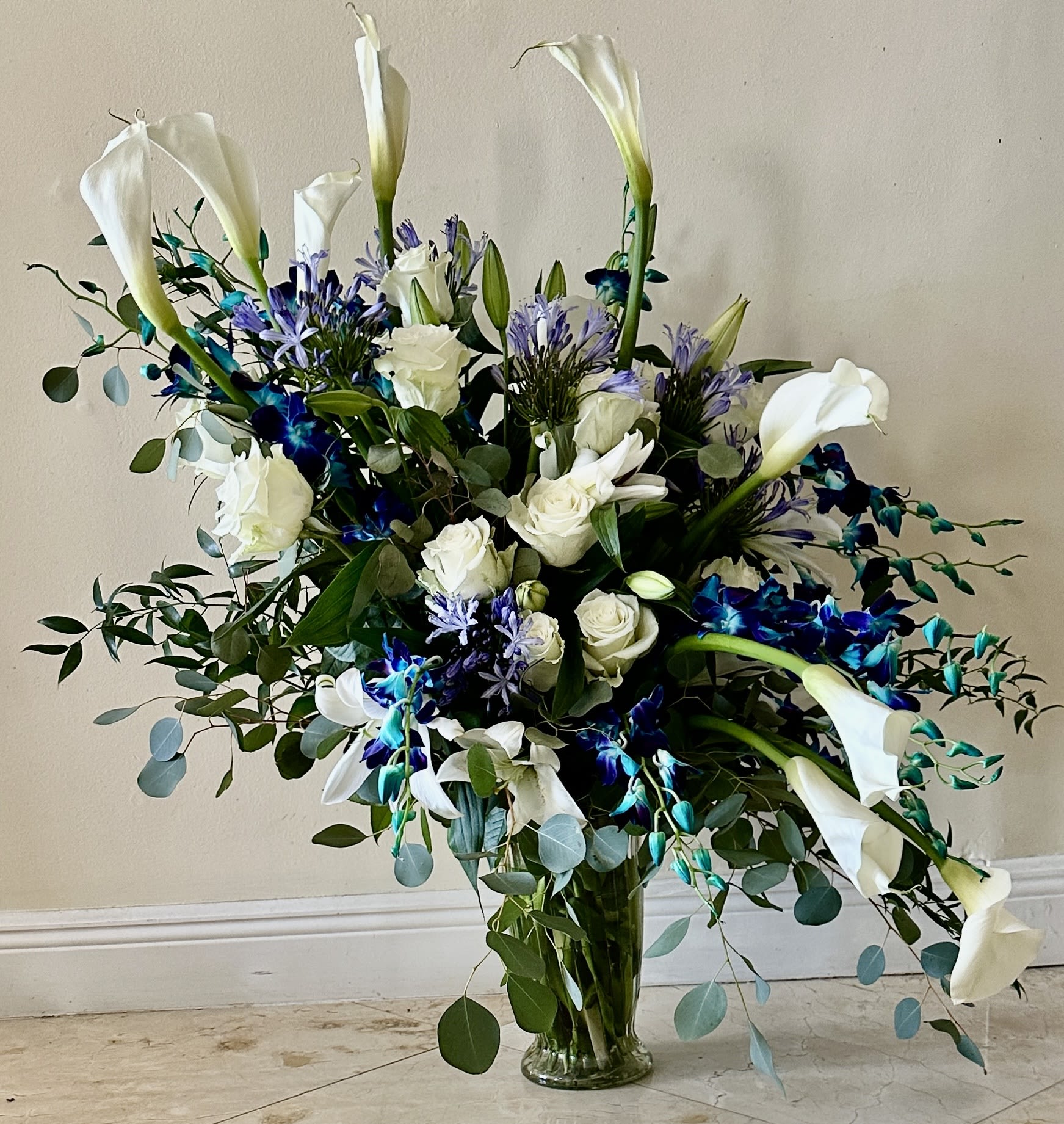 Tall bouquet of white calla lilies, roses, and blue flowers in a glass vase