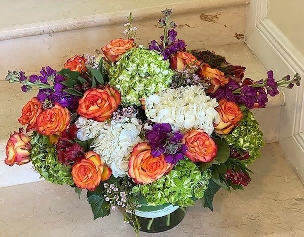 Low round arrangement of orange roses, green and white hydrangeas, and purple blooms in a glass vase