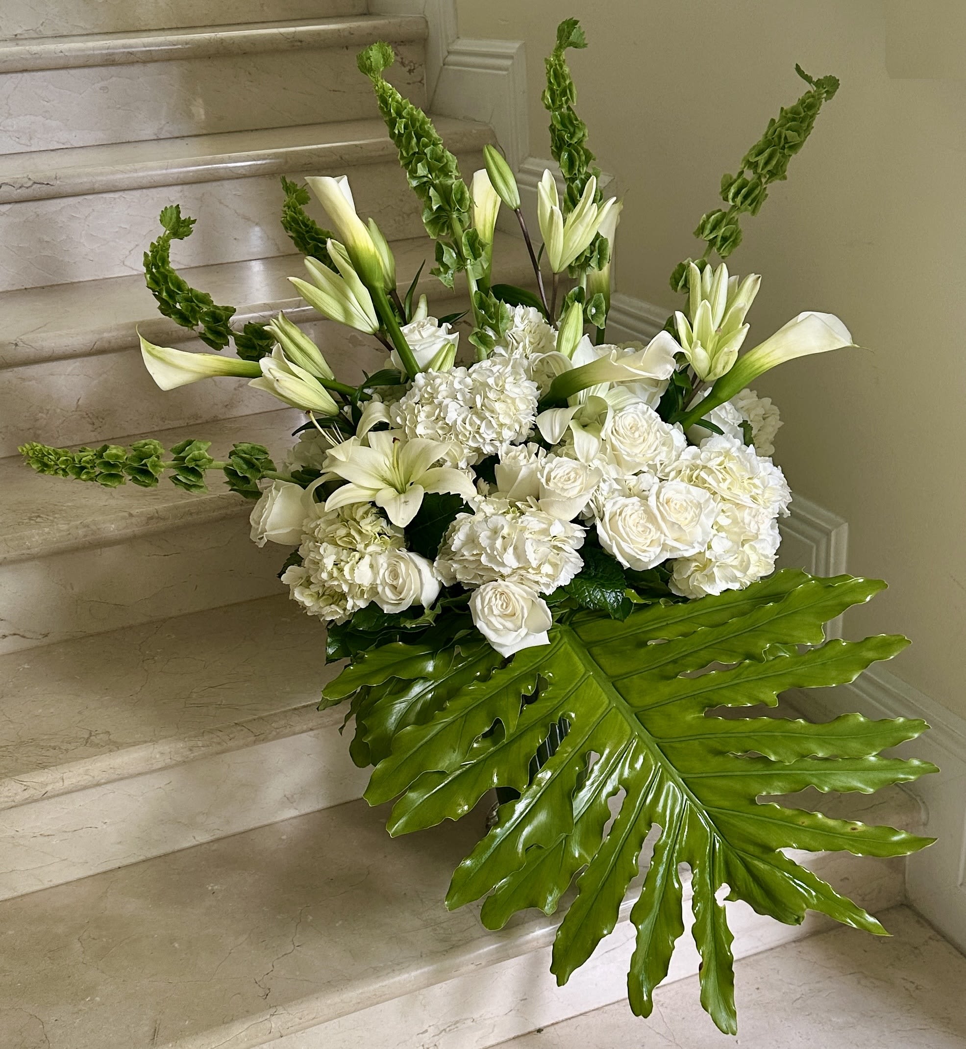 Large white arrangement with lilies, roses, and a big green tropical leaf on marble steps