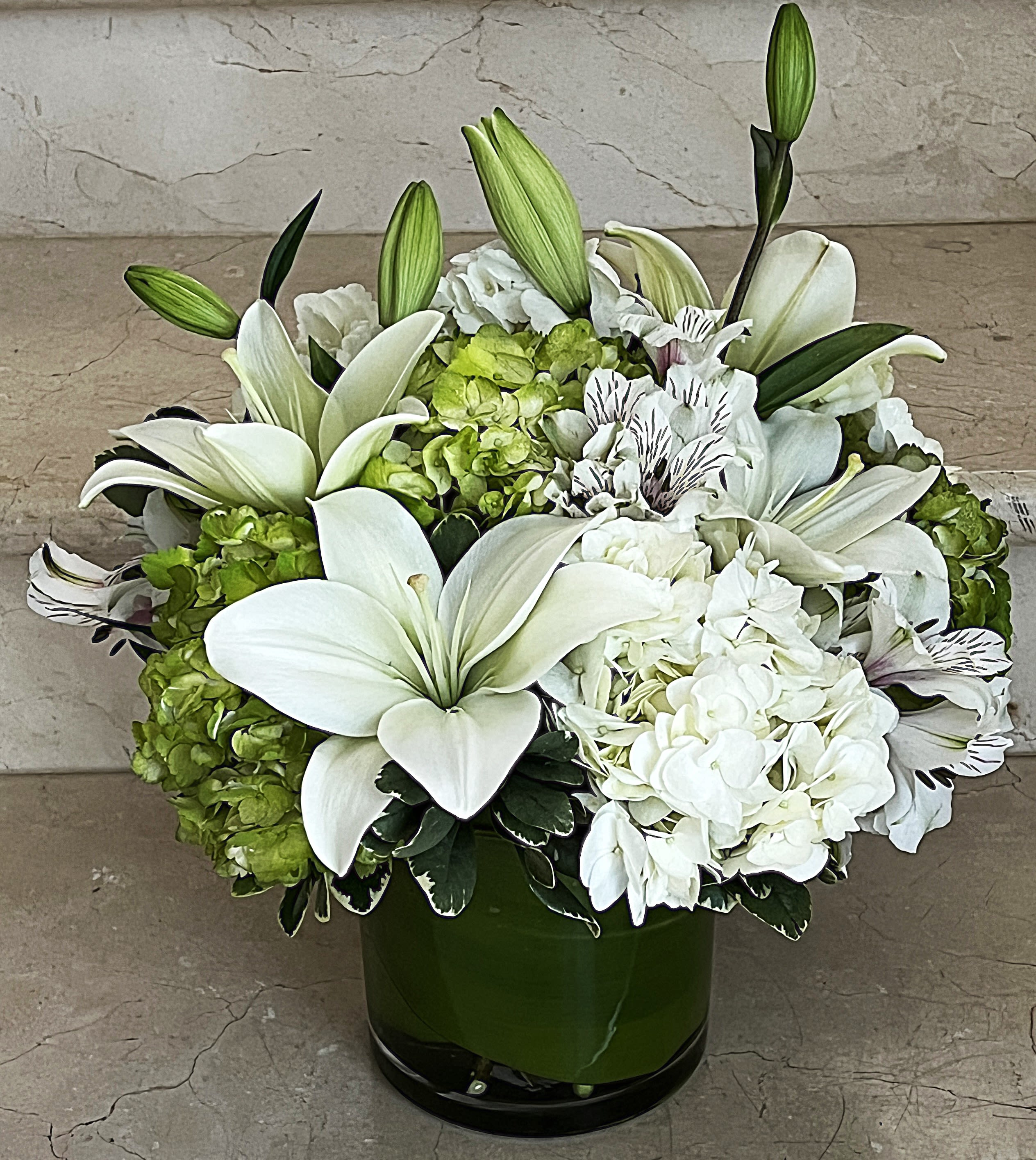 Low arrangement of white lilies, hydrangeas, and alstroemeria in a green glass cylinder vase