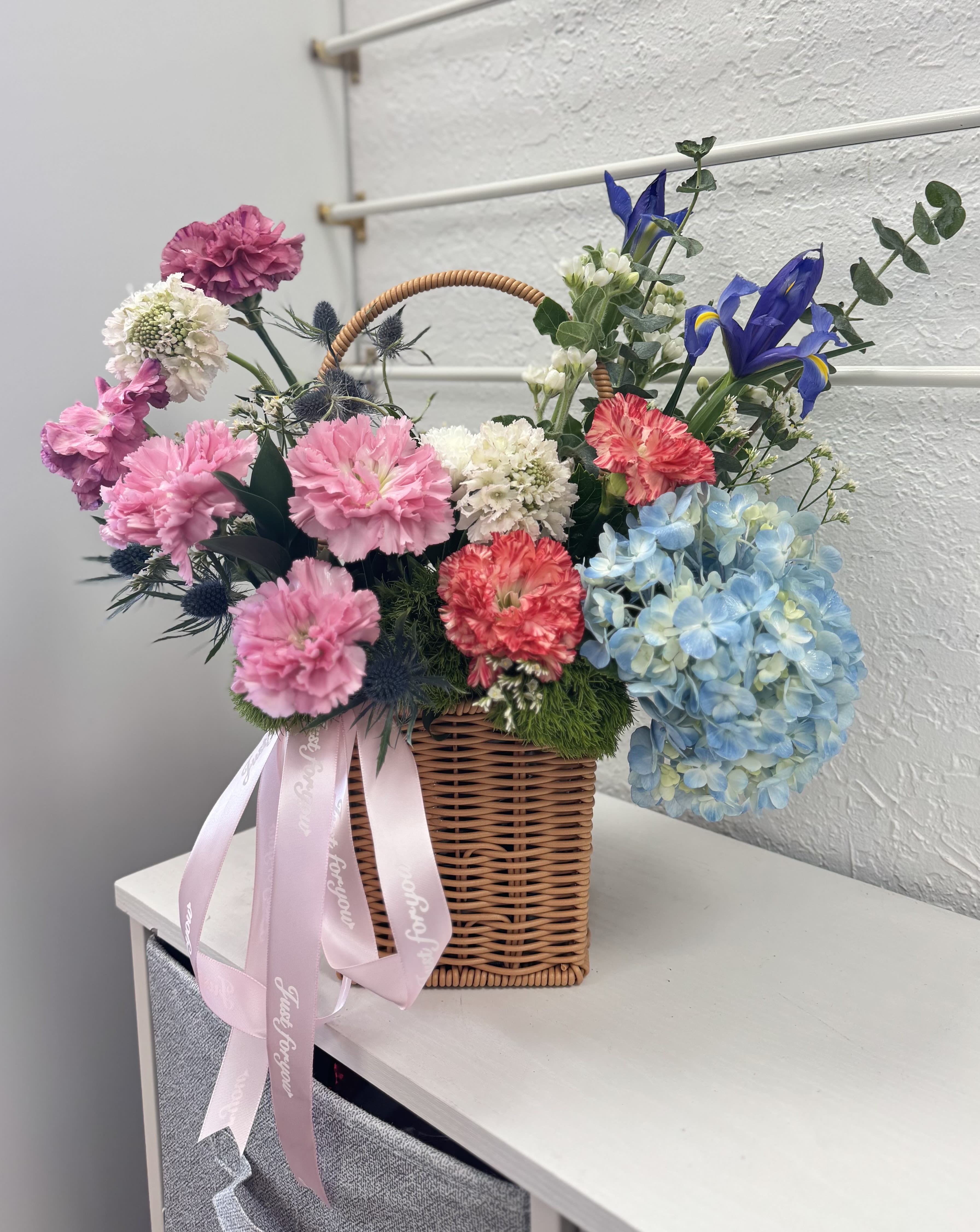 Basket arrangement of pink, blue, and white flowers with a pink ribbon