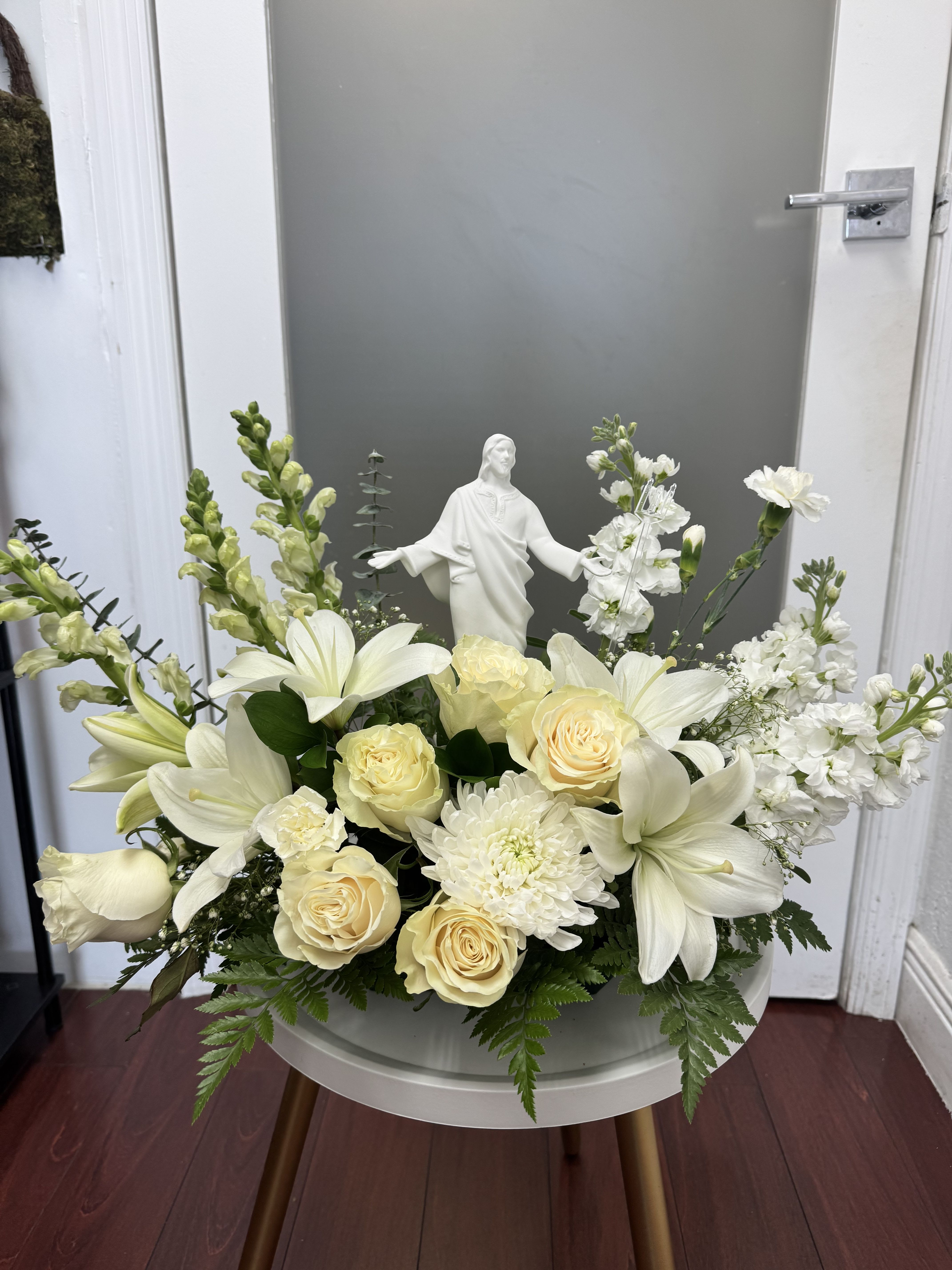 White floral arrangement with roses, lilies, and a small religious statue