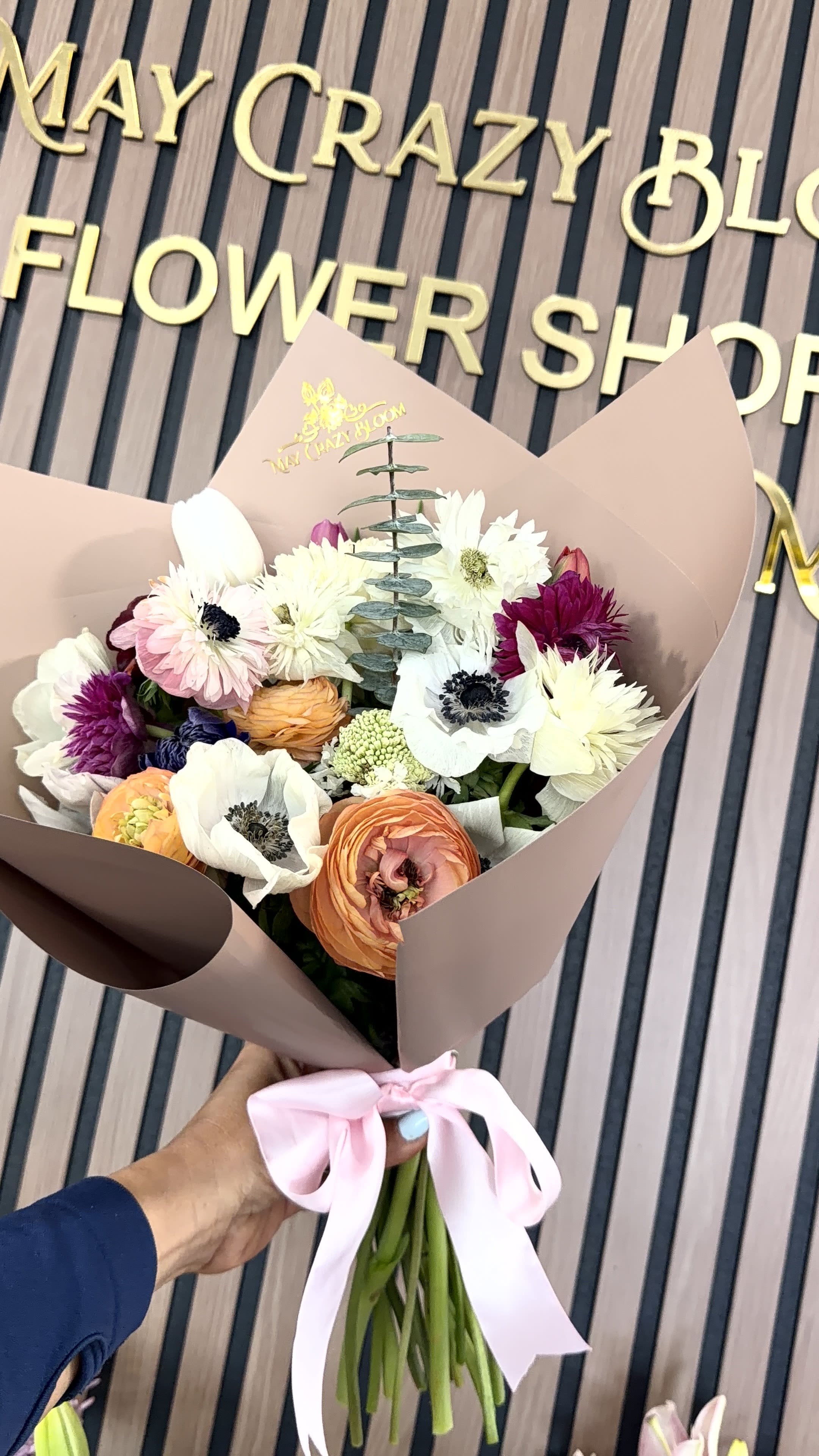 Anemone Story  - A rounded hand-tied bouquet of anemone, ranunculus, tulips and daisy-like blooms in a cheerful mix of white, blush pink, purple and lively orange-Anemone Story is the kind of birthday bouquet or anniversary arrangement that reads as both romantic and joyful. The dark-centered anemones and layered ranunculus draw the eye, while pops of orange and soft tulip curves keep the composition bright and modern; it's an arrangement that makes the person you're thinking of feel seen and celebrated.  This bouquet's dense, layered texture pairs jewel-toned petals with soft pastels for contrast and depth. Tall eucalyptus stems add a graceful vertical accent through the rounded cluster; the stems are finished in blush paper wrap and tied with pale pink satin and ribbon for a polished, gift-ready presentation. Perfect as a thank-you gift, just-because surprise, or everyday celebratory bouquet, Anemone Story arrives ready to display and brighten any room.