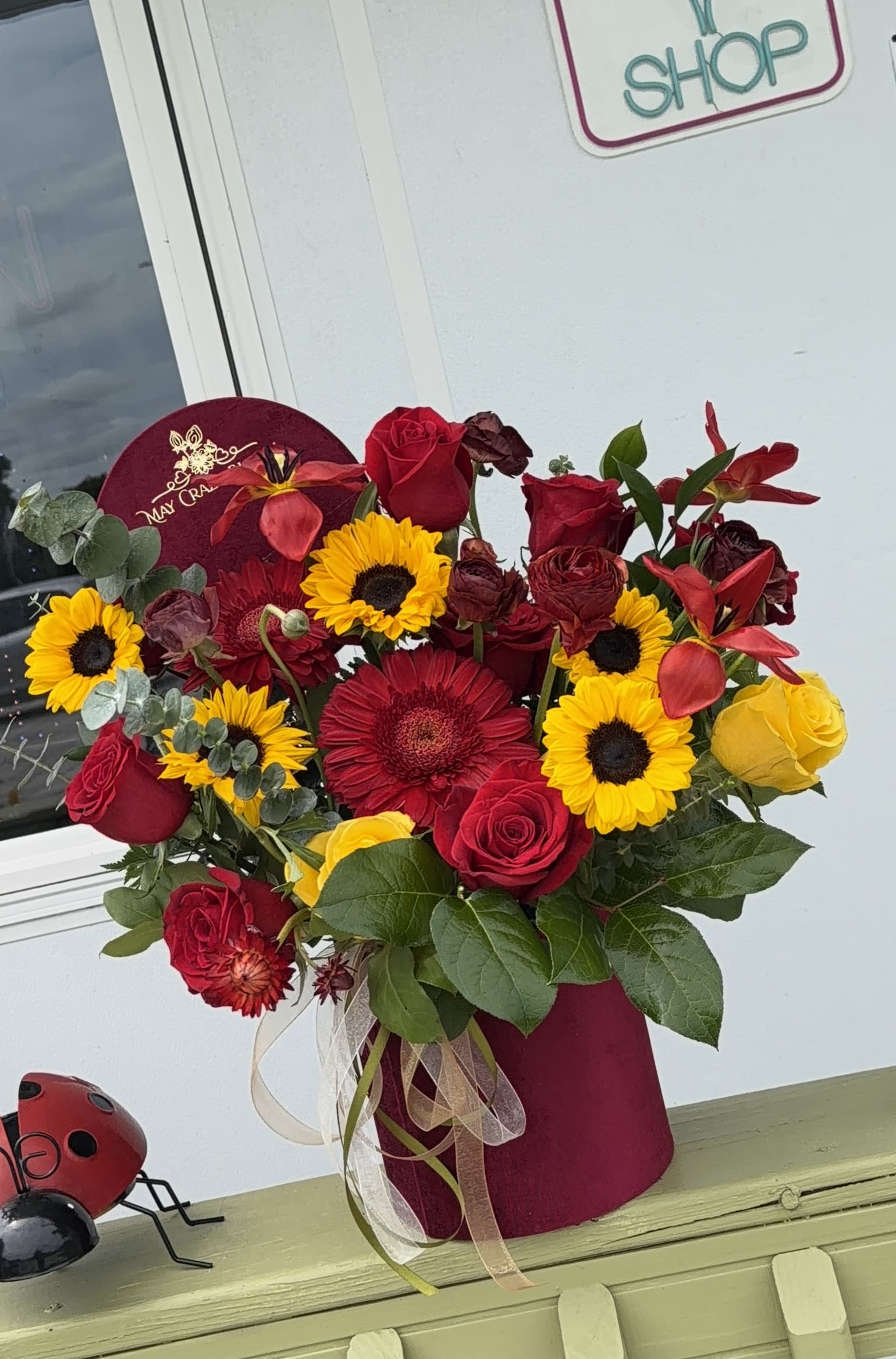 Red and Yellow Priceless - A bold, high-contrast arrangement that makes someone feel celebrated the moment they see it - perfect as a birthday bouquet, anniversary roses substitute, or a festive surprise. Deep red roses and a large red gerbera daisy pair with bright yellow sunflowers to create warm, striking color play; the combination reads both romantic and joyful so the person you're sending it to will feel noticed and cherished.  This lush arrangement is presented in a rounded burgundy hat box for an elegant, gift-ready look. Smaller burgundy blooms and red accent flowers add depth and texture while green foliage lifts the composition; a clear ribbon at the base and a round floral pick finish the design. Sized to fill the hat box with abundant, garden-fresh presence, it arrives ready to display and brighten a table, entryway, or office - no vase needed.