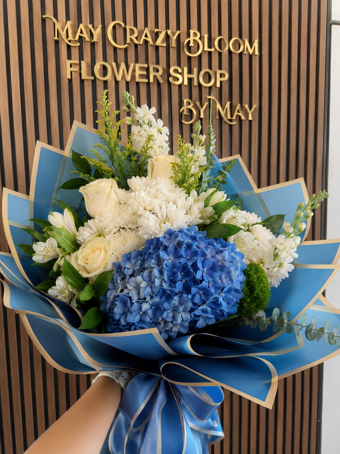 Blue Universe - A striking cluster of blue hydrangea sets the tone for this blue-and-white flower arrangement-perfect as a birthday bouquet, congratulations gift, or a spring celebration bouquet that feels both fresh and refined. The cool blue and soft white palette reads calm and elegant, the kind of gift that makes the person you're thinking of feel noticed and quietly celebrated.  Creamy white roses and full white chrysanthemums surround the hydrangea, with delicate white accent blooms and alstroemeria adding texture and movement against fresh green foliage. The bouquet is wrapped in layered blue paper with subtle beige edging and finished with a wide blue satin ribbon for a polished, gift-ready presentation that photographs beautifully and arrives secure.  Choose this arrangement when you want to say thank you, congratulations, or happy birthday with a graceful, slightly formal touch-ideal for a colleague, mother, or close friend who appreciates cool tones and classic blooms. Standard size fits comfortably in most vases; to keep flowers looking their best, recut stems and change the water every 2-3 days.