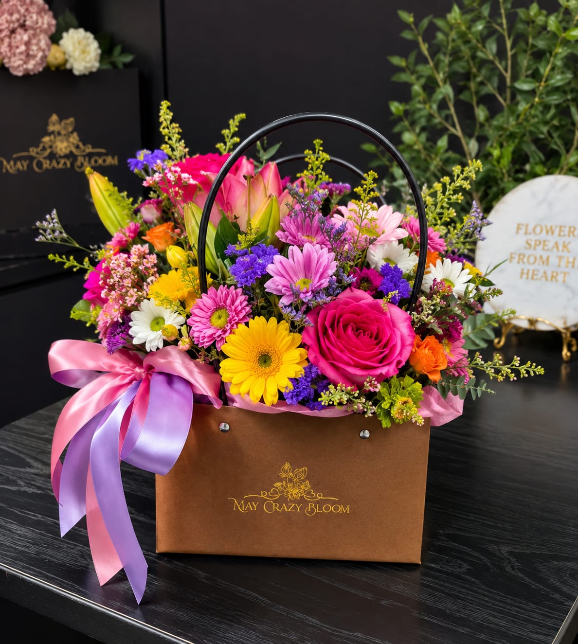 Late afternoon  - Bright pink roses and lively gerbera-style daisies spill from a brown gift bag tied with pink and lavender satin ribbons, creating a cheerful late-afternoon glow perfect for a birthday bouquet or a thank-you arrangement. This flower arrangement pairs vibrant pinks and purples with sunny yellow and crisp white blooms to deliver an expressive, celebratory impression-the kind that makes the person you're thinking of feel noticed and genuinely cheered.  The mixed bouquet features bright pink roses, yellow and pink daisy-like blooms, white daisies, orange accents, purple filler flowers, and a few lily buds and open lily blooms for added height and graceful texture. Everything is arranged in a brown handled gift bag with a black handle and finished with large pink and purple satin ribbons, giving the display a polished, gift-ready presentation ideal for springtime occasions or everyday celebrations.  Perfect for sending to a friend, coworker, or family member who deserves a burst of color, this arrangement reads as joyful and personal without being overdone. Order with standard sizing and expect a layered, hand-tied composition that arrives ready to display-no extra vase required for the immediate gifting moment.