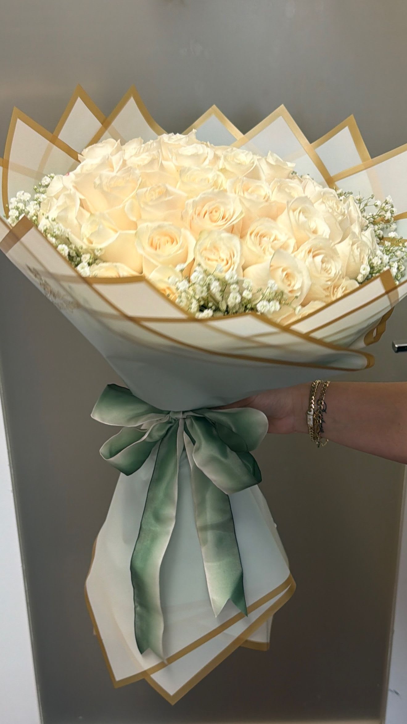 100 wrap white roses  - A lush dome of one hundred cream-to-white roses creates an unforgettable birthday bouquet, anniversary surprise, or elegant sympathy arrangement-the kind of floral gift that makes the person you're thinking of feel deeply seen. The predominantly white blooms carry soft hints of blush and peach in a few petals, lending warmth and subtle dimension that reads romantic for Valentine's Day or quietly reverent for Memorial Day.  Tightly packed for a full, rounded silhouette, the roses are finished in crisp white paper wrap with delicate gold edging and a decorative outer wrap, then tied with a soft green satin ribbon for a polished, gift-ready presentation. Small white filler blooms peek at the edges to soften the shape and enhance the bouquet's texture; the result is refined, tactile, and photo-ready.  Perfect for Thanksgiving hostess gifts, Women's Day tributes, or a major milestone moment, this arrangement arrives ready to impress and comfort. For best longevity, keep stems in fresh water, trim stems slightly before placing in a clean vase, and display away from direct heat or drafts to extend the life of the roses.
