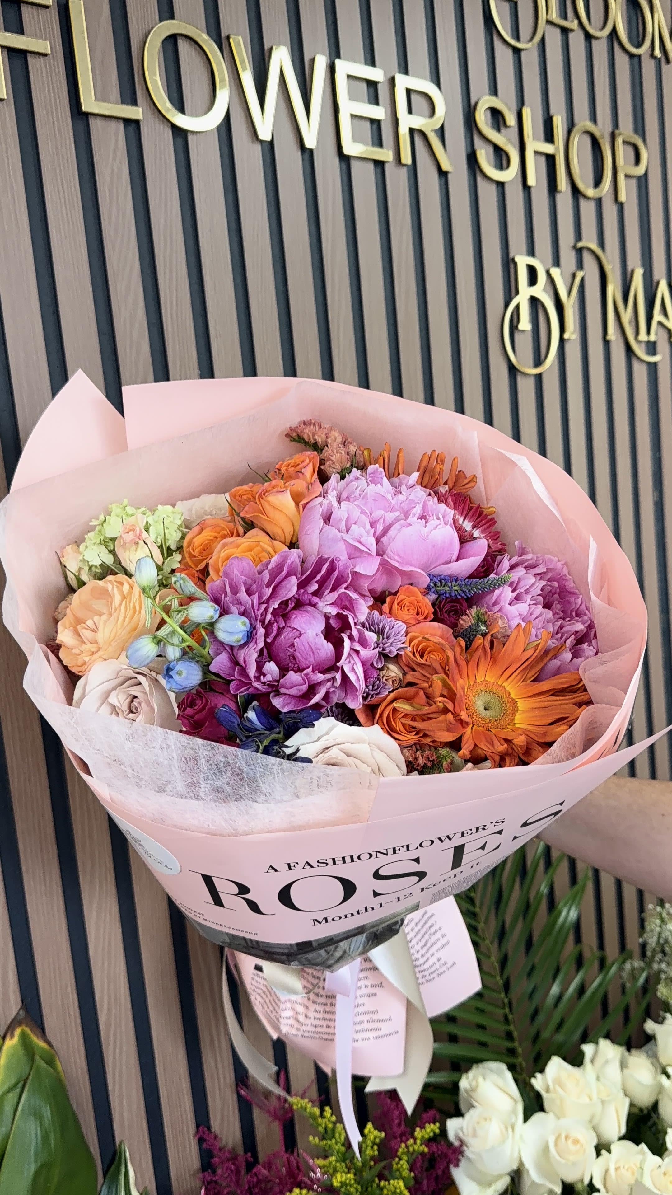 Caos en mi Universo  - A lively, layered bouquet bursting with springtime joy-this hand-tied flower arrangement is perfect as a birthday bouquet or cheerful celebration gift. Bright pink and soft lavender peony-like blooms mingle with clustered roses and a striking orange gerbera daisy to create an energetic, romantic look that will make the person you're thinking of feel truly celebrated.  The mix includes roses, peony-style blooms, gerbera daisy, hydrangea touches and smaller daisies and accents in a palette of pink, orange, white and purple. Arranged densely for a full, textured silhouette, the stems are finished in pale pink paper wrap and tied with ribbon for a polished, gift-ready presentation. The bouquet reads fresh and playful at table-top size-designed to stand out in photos and fill a room with color.  Ideal for birthdays, springtime occasions, or to send someone a bright pick-me-up, this bouquet's cheerful colors suit recipients who love vibrant, romantic arrangements. For best longevity, keep stems in fresh water, trim about an inch before placing in a clean vase, and display away from direct sun and heat.