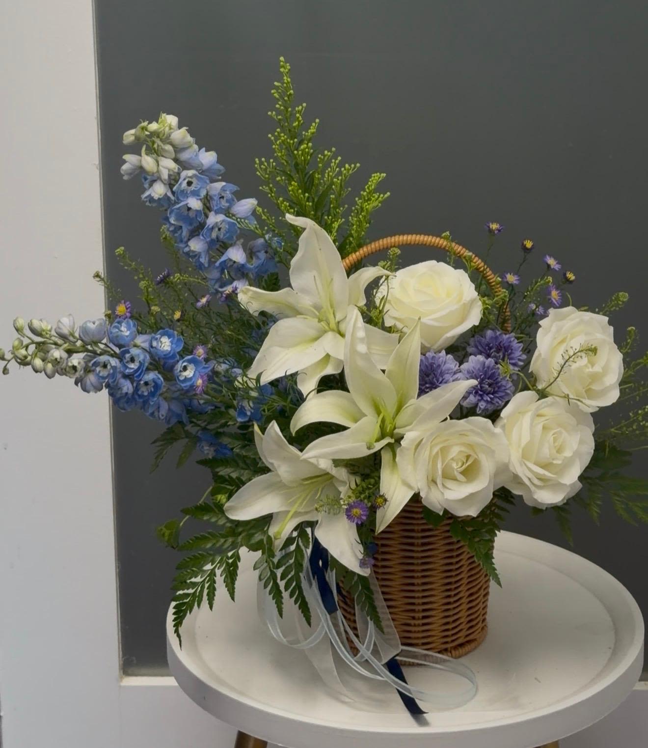 Night Whispers  - Cool, elegant and quietly dramatic-this basket arrangement brings together white lilies and cream roses with tall blue delphinium spikes for a look that feels both refined and comforting. As a sympathy arrangement, thinking-of-you gift, or a tasteful birthday bouquet, Night Whispers says you care with calm confidence and fresh, sophisticated color.  Arranged in a woven wicker basket, the design pairs large white lilies and soft cream roses with airy greenery and small white filler blooms to create a layered, full silhouette. The blue, delphinium-like spikes add vertical height and a serene contrast against the ivory tones, while the wicker base gives the composition a warm, natural touch that suits home or bedside display.  Perfect for the person who prefers understated elegance, this arrangement conveys sympathy, gratitude, or quiet celebration without overwhelming. Sizes are designed for easy tabletop placement; to keep blooms looking their best, refresh the water daily, remove any spent flowers, and keep the basket away from direct heat and drafts.