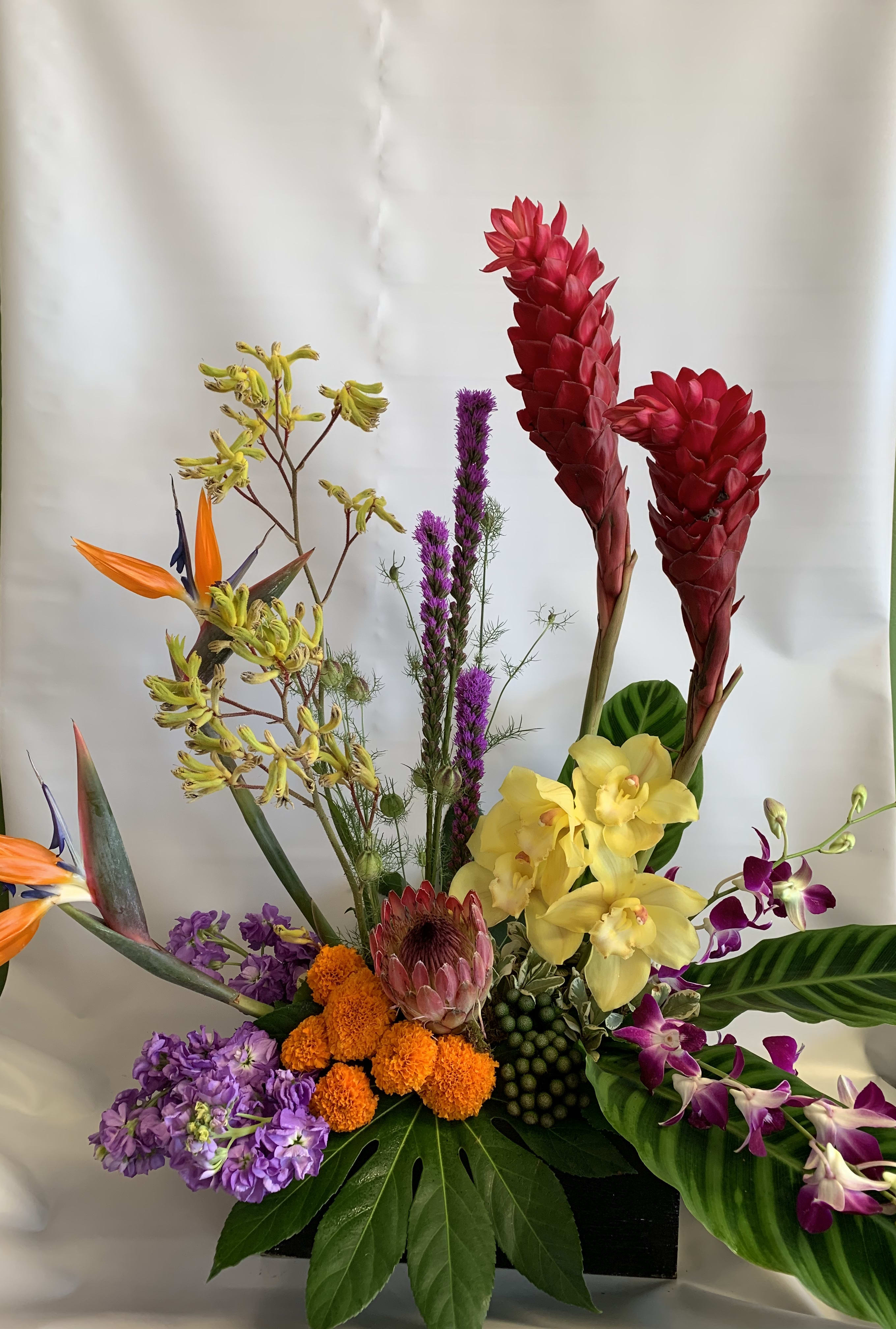 Tropical floral arrangement with red ginger, yellow orchids, and purple blooms in a black container
