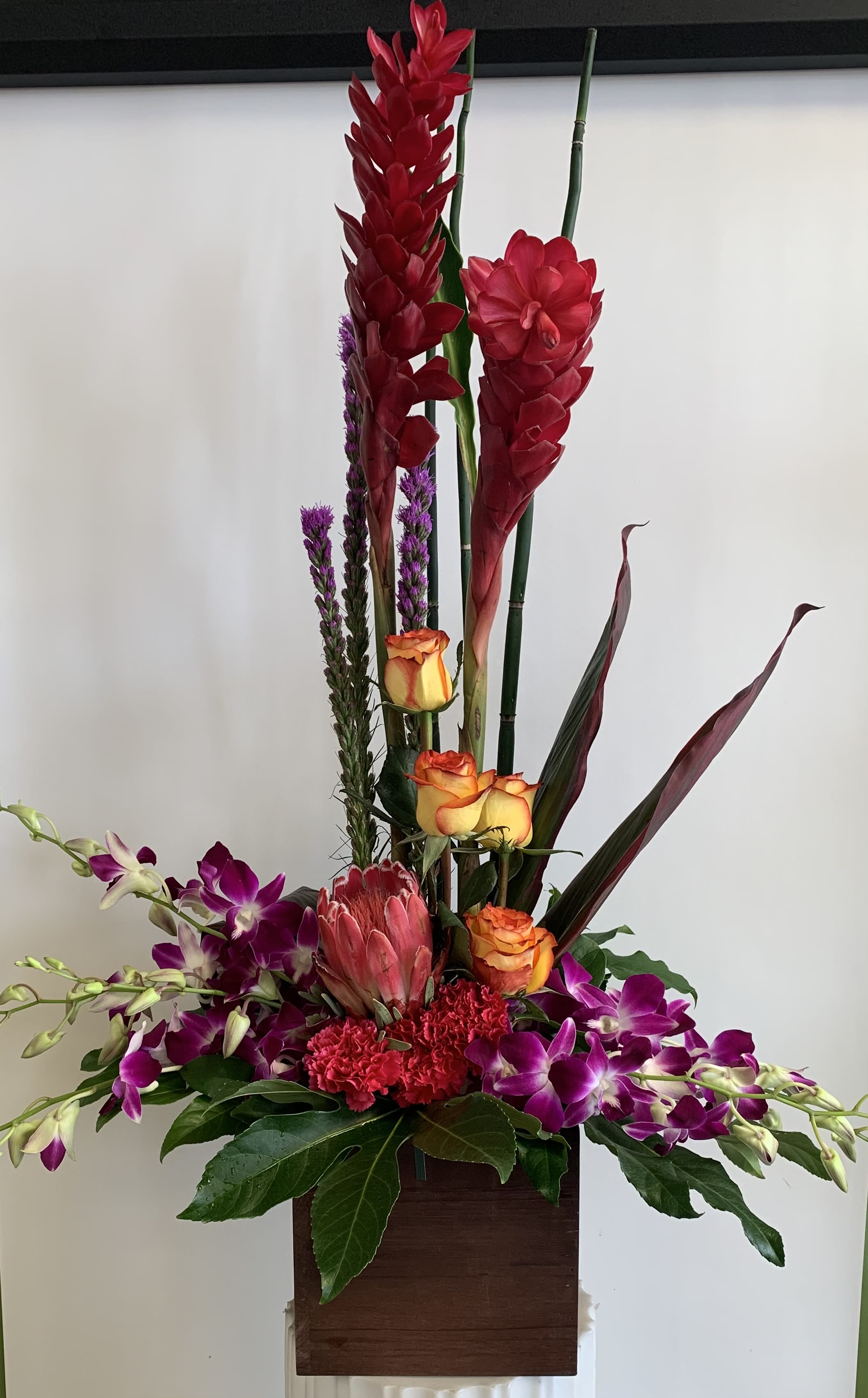 Tall tropical arrangement with red blooms, orange roses, and purple orchids in a wooden container