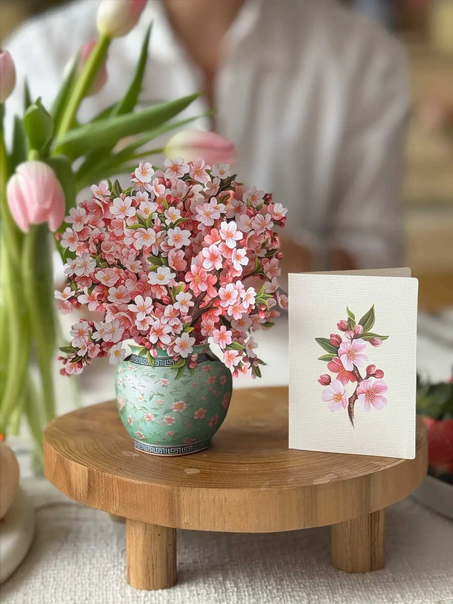 Pink cherry blossom arrangement in a green patterned vase beside a matching card