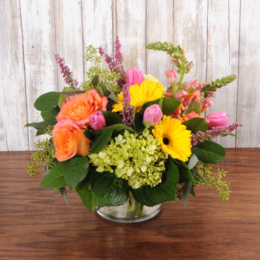 Low glass vase arrangement with orange roses, yellow gerbera daisies, pink tulips, and green hydrangea.