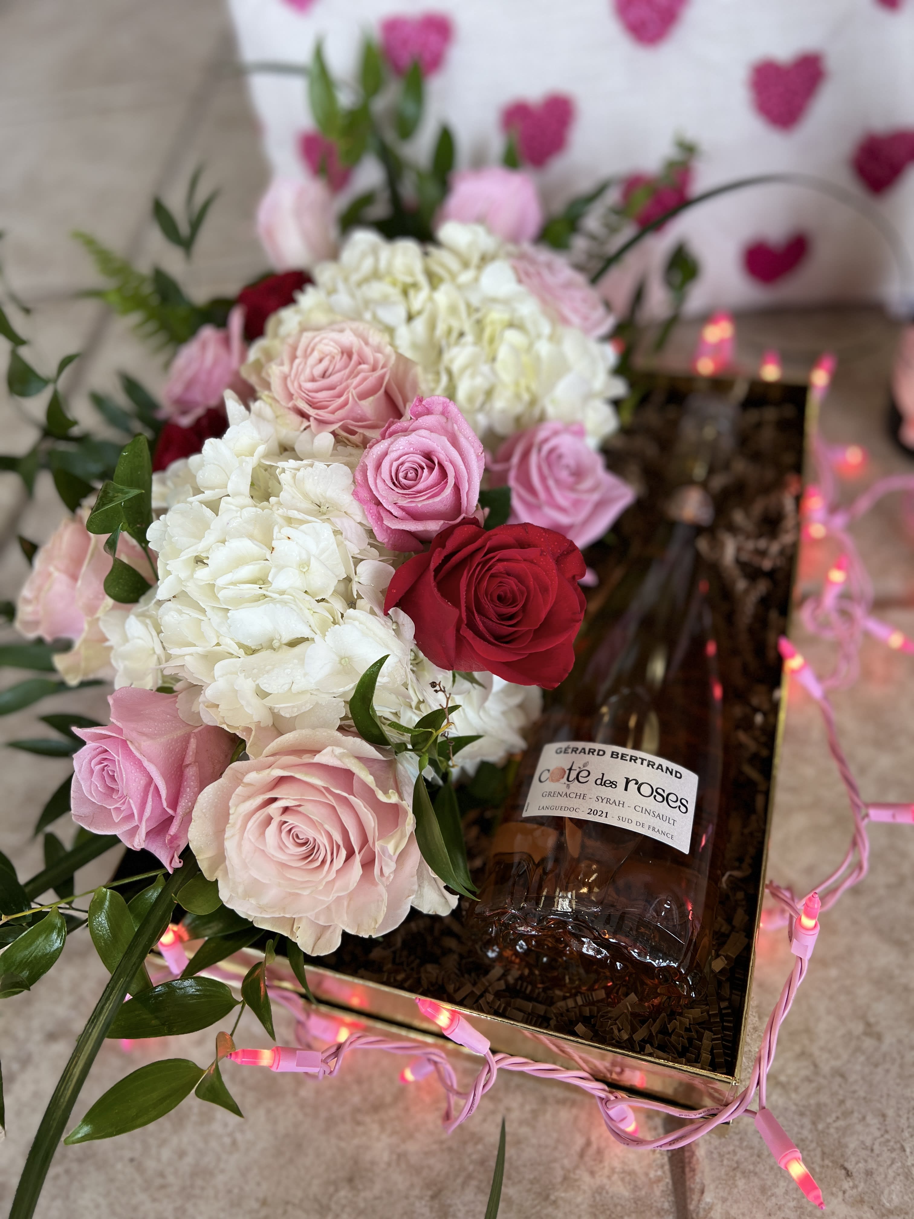 Pink and red roses with white hydrangeas in a gift box beside a wine bottle