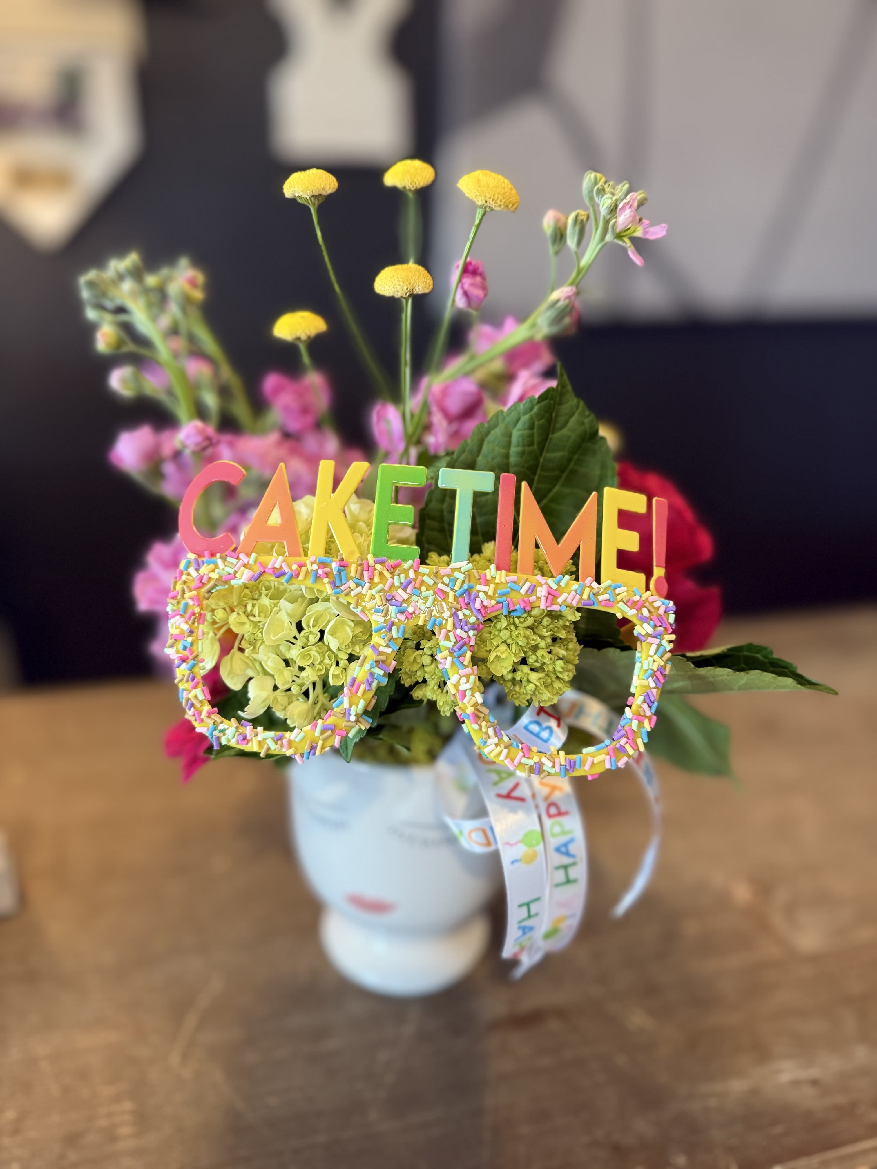 Flower arrangement in a white mug with a colorful "CAKE TIME!" topper