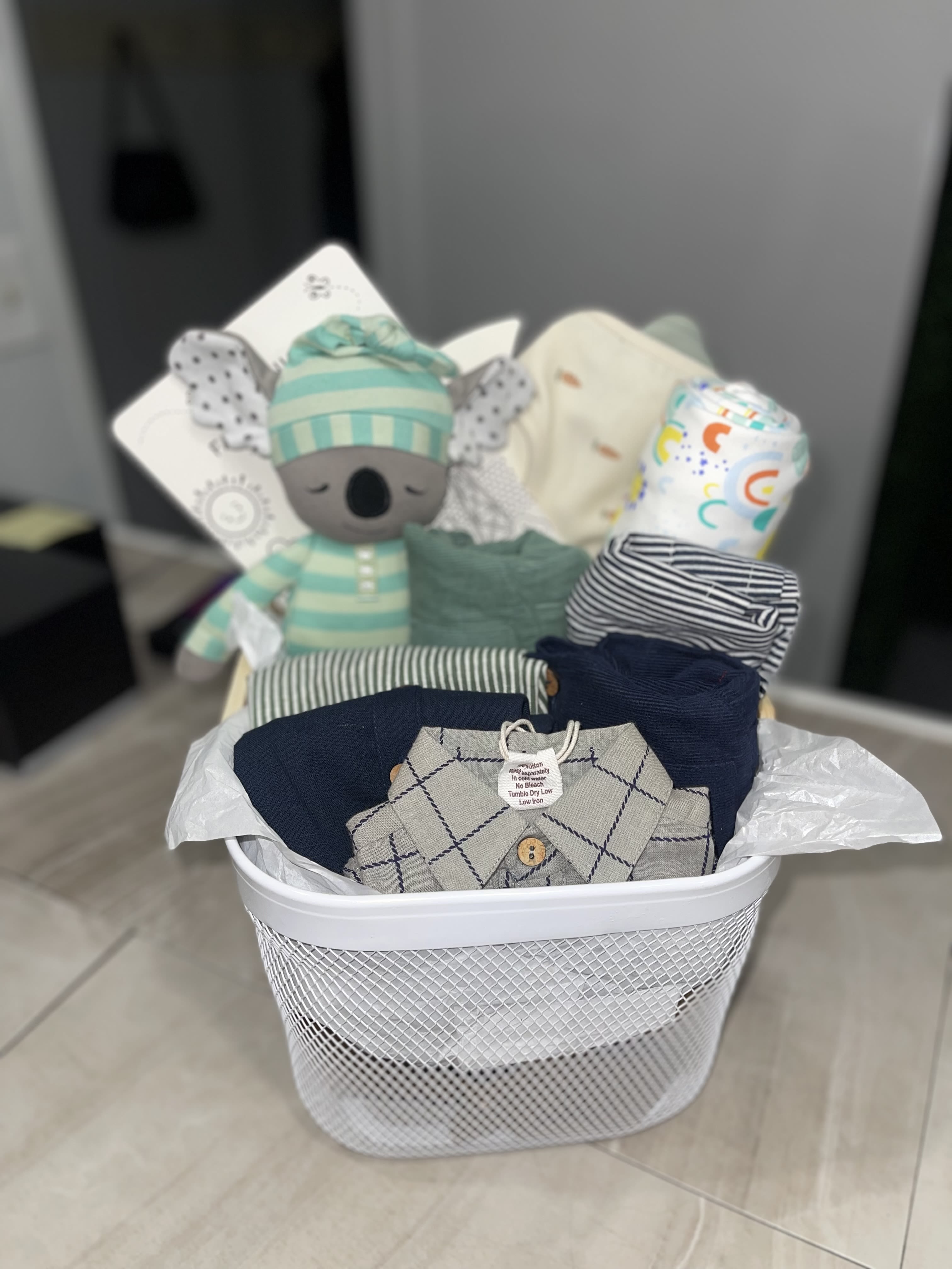 Medium Hello Baby Boy Basket - Soft neutrals washed with navy, mint and pastel accents make this baby gift basket feel like a cozy nursery hug - perfect as a new baby or baby shower gift for a baby boy. The plush koala and other cuddly toys give the recipient a playful focal point, while the white mesh or woven laundry-style basket presents everything neatly for a welcome‑home moment that says "we're so glad you're here."  Inside you'll find folded infant clothing in neutral, striped and blue tones, This basket is made to be given: thoughtful for new parents, sweet for a shower, and useful every day afterward. Choose the white mesh laundry-style option or woven basket presentation at checkout; items arrive gift-wrapped with a tag so the little one's first cuddles start the moment it's opened.