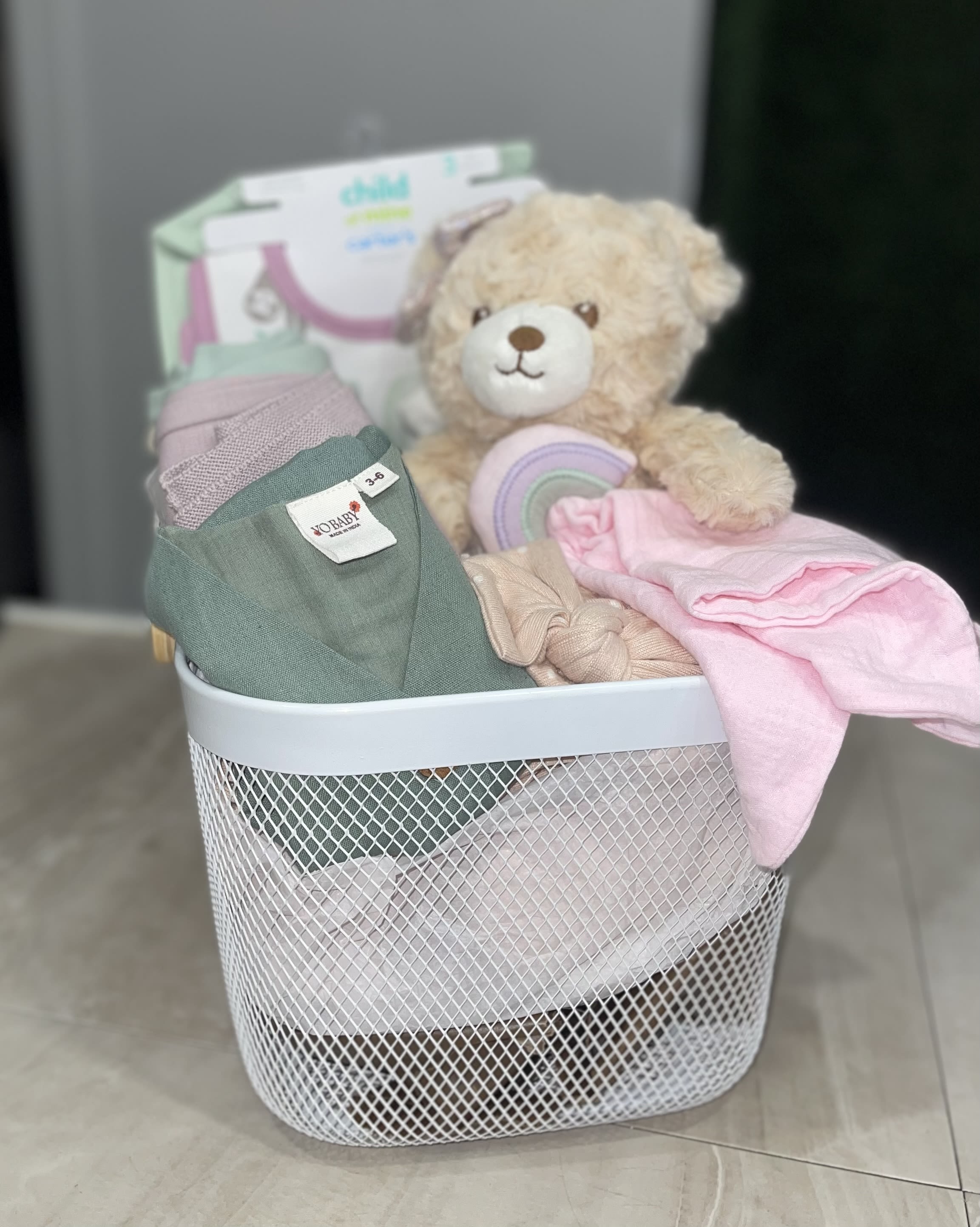 Medium Hello Baby Girl Basket - Soft pastels and cuddly textures make this Hello Baby Girl Basket the kind of newborn gift that instantly comforts and delights. Perfect as a new baby or baby shower present, the baby gift basket reads like a thoughtful welcome-home surprise for the little girl you're celebrating - she'll be surrounded by soft blankets, plush friends, and sweet essentials that set a gentle, nursery-ready tone.  Arranged upright in a white wire  basket, this set includes a plush teddy with folded baby clothing and blankets in pink, mint, lavender, packaged accessories. The layered presentation and mixed textures - soft knits, cotton garments, - create a tidy, gift-ready look that's easy to place in the nursery.   This basket is ideal for the new mom who could use cozy essentials and a little pampering; it's the kind of present that says "congratulations" while helping baby feel snug and loved. 
