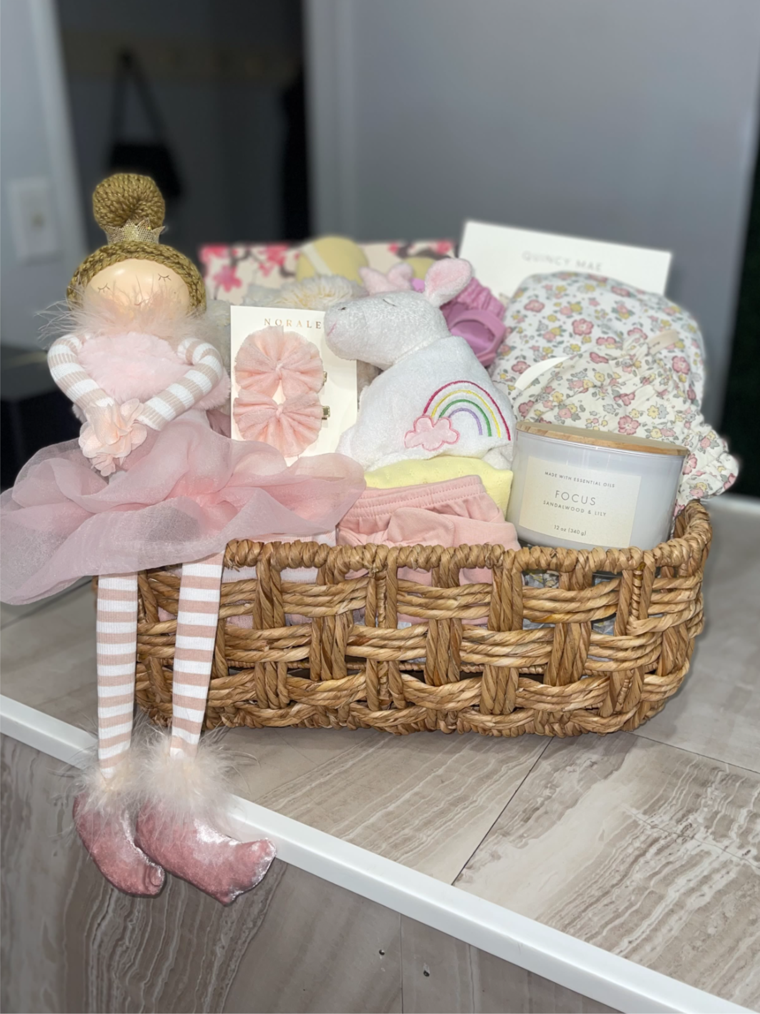 Large Hello Baby Girl Basket - Soft pastels and cuddly textures make this Hello Baby Girl Basket the kind of newborn gift that instantly comforts and delights. Perfect as a new baby or baby shower present, the baby gift basket reads like a thoughtful welcome-home surprise for the little girl you're celebrating - she'll be surrounded by soft blankets, plush friends, and sweet essentials that set a gentle, nursery-ready tone.  Arranged upright in a white wire or woven basket, this set includes a plush teddy or doll, folded baby clothing and blankets in pink, mint, lavender, peach and cream, plus curated extras like a scented candle jar, packaged accessories. The layered presentation and mixed textures - soft knits, cotton garments, and wicker basket - create a tidy, gift-ready look that's easy to place in the nursery. And A variety of mixed blooms and foliage in a glass bud vase.  This basket is ideal for the new mom who could use cozy essentials and a little pampering; it's the kind of present that says "congratulations" while helping baby feel snug and loved. 