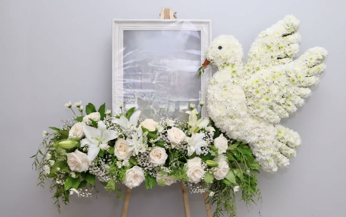 White floral funeral spray with roses and lilies beside a framed tribute