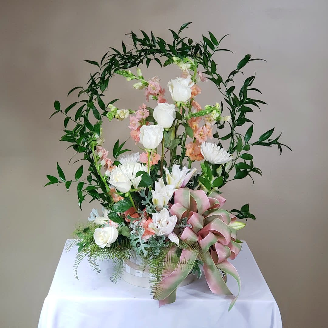 Floral arrangement with white roses and lilies in a basket, framed by greenery and a pink ribbon