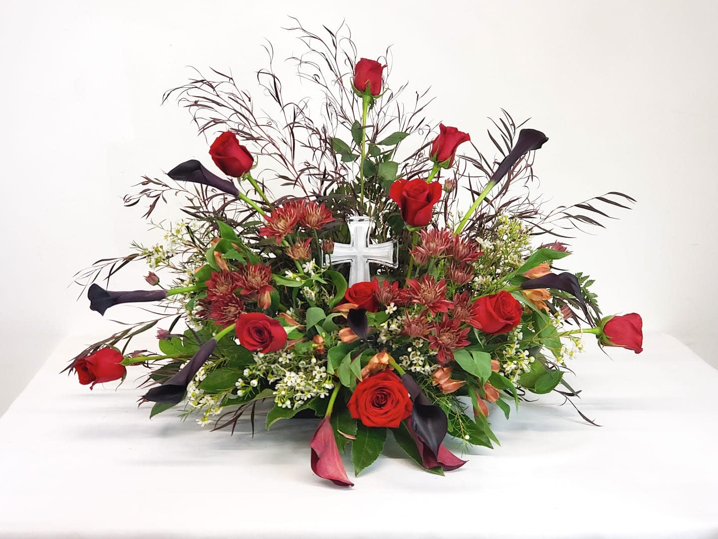 Red roses and dark calla lilies in a low floral arrangement with a white cross