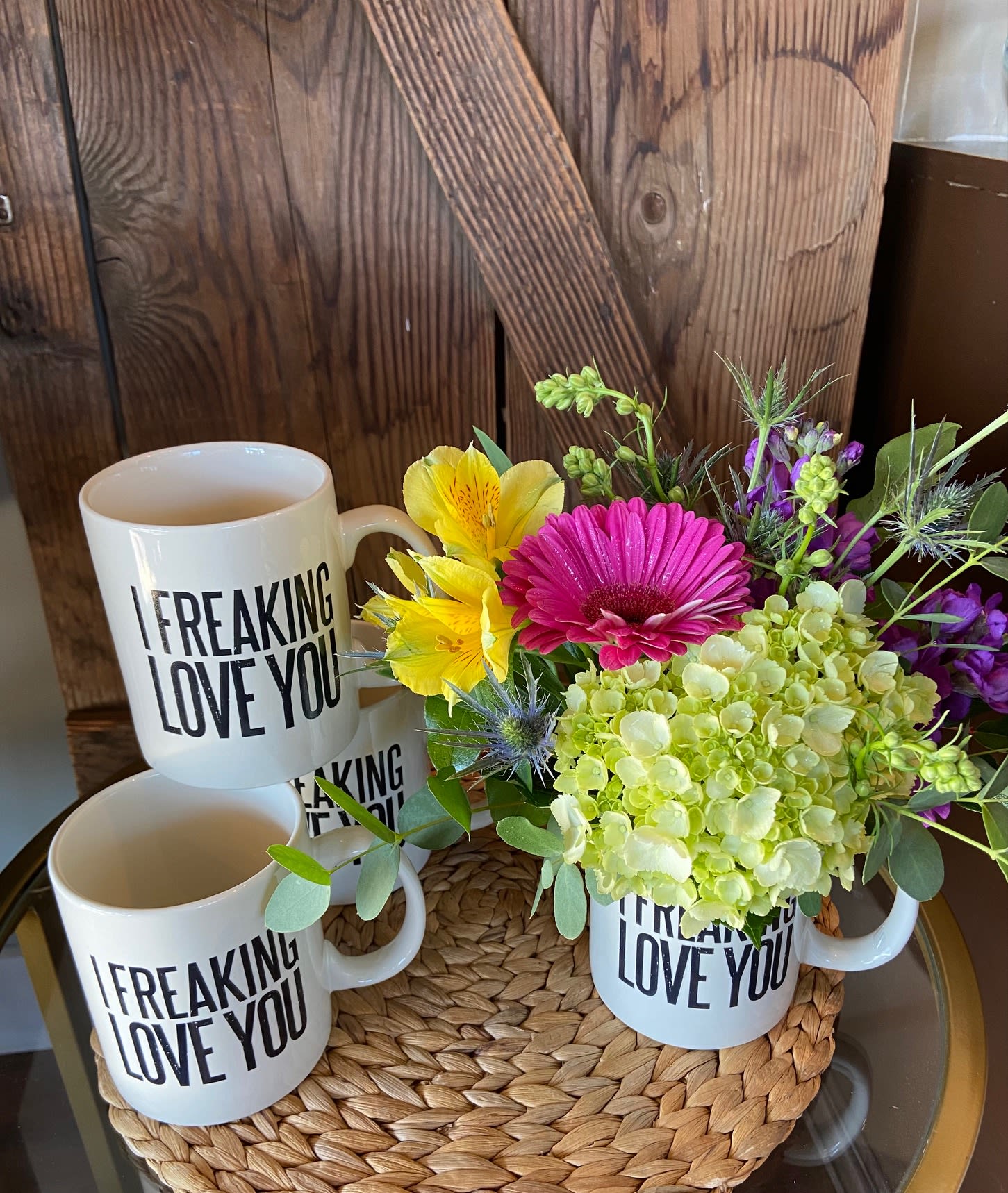 White 'I freaking love you' mugs, one filled with pink, yellow, and green flowers on a woven mat.