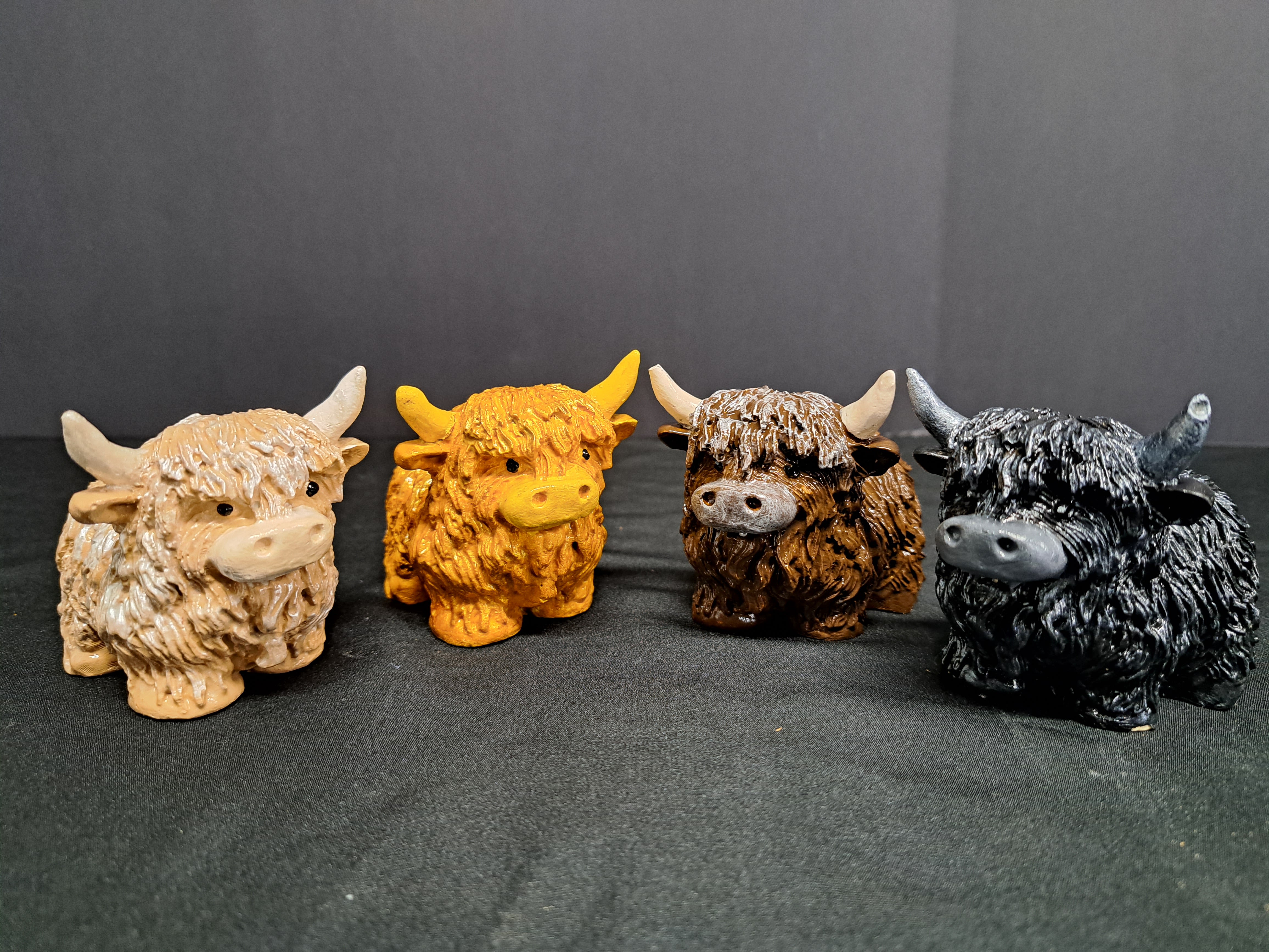 Highland Cows "Coos" - Playful collectible accent pieces shaped like Highland cows bring a touch of rustic charm to gift-giving and home décor. This mixed-color set of four small cow figurines-tan, bright yellow, brown, and black-reads as a whimsical gift for birthdays, housewarmings, or anyone who loves Scottish-inspired accents. The row of shaggy, textured figures makes a cheerful tabletop display that feels personal and lighthearted.  Each ceramic figurine has a textured, shaggy finish that catches the light and invites a closer look; the varied colors create a lively, cottage-style grouping perfect for a mantel, shelf, or desk. Sold individually or as a set, these compact decorative cows are easy to place and pair well with seasonal florals or as a standalone keepsake. They require no special care-just dust lightly-making them an effortless way to make the person you're thinking of smile.