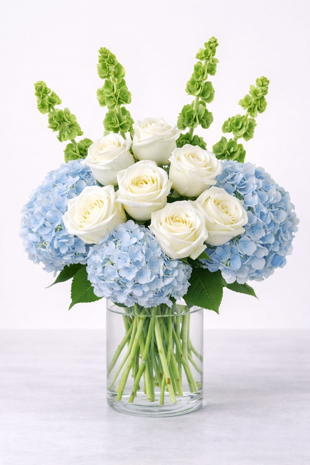 White roses and blue hydrangeas in a clear glass vase with green spikes