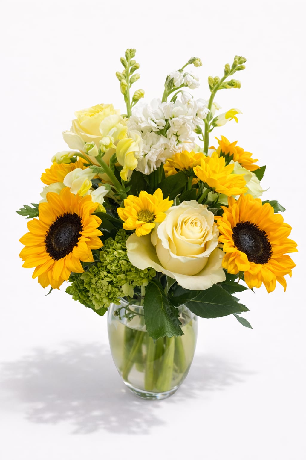 Golden Smile Bouquet - Bright, joyful, and full of life, this charming floral arrangement brings together radiant sunflowers, elegant ivory roses, soft yellow blooms, and lush seasonal greenery in a clear glass vase.  The golden sunflowers symbolize happiness and warmth, while the delicate roses add a touch of romance and elegance. Together they create a cheerful design that instantly brightens any room and lifts the spirit.  Fresh, vibrant, and filled with sunshine — this arrangement is the perfect way to celebrate birthdays, send encouragement, or simply make someone smile.