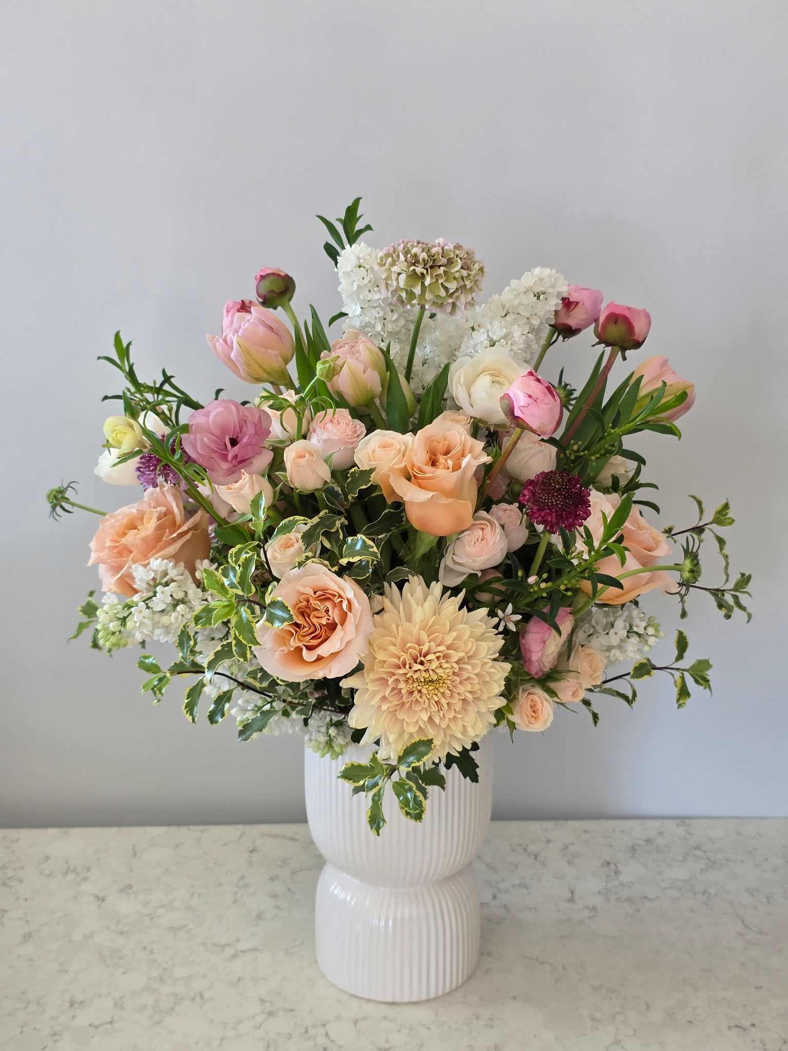 Sweet Darling  - Sweet Darling is a romantic garden-style arrangement designed to celebrate love in its most graceful form. Featuring a lush blend of soft peach roses, lush pink peonies, delicate blush ranunculus, creamy white blooms, and seasonal accents, this piece is artfully arranged in a modern ribbed ceramic vase.  Layers of texture—from airy florals to rich focal blooms—create a full, dimensional design that feels both refined and effortlessly natural. Perfect for anniversaries, this arrangement expresses warmth, admiration, and enduring love with every petal.