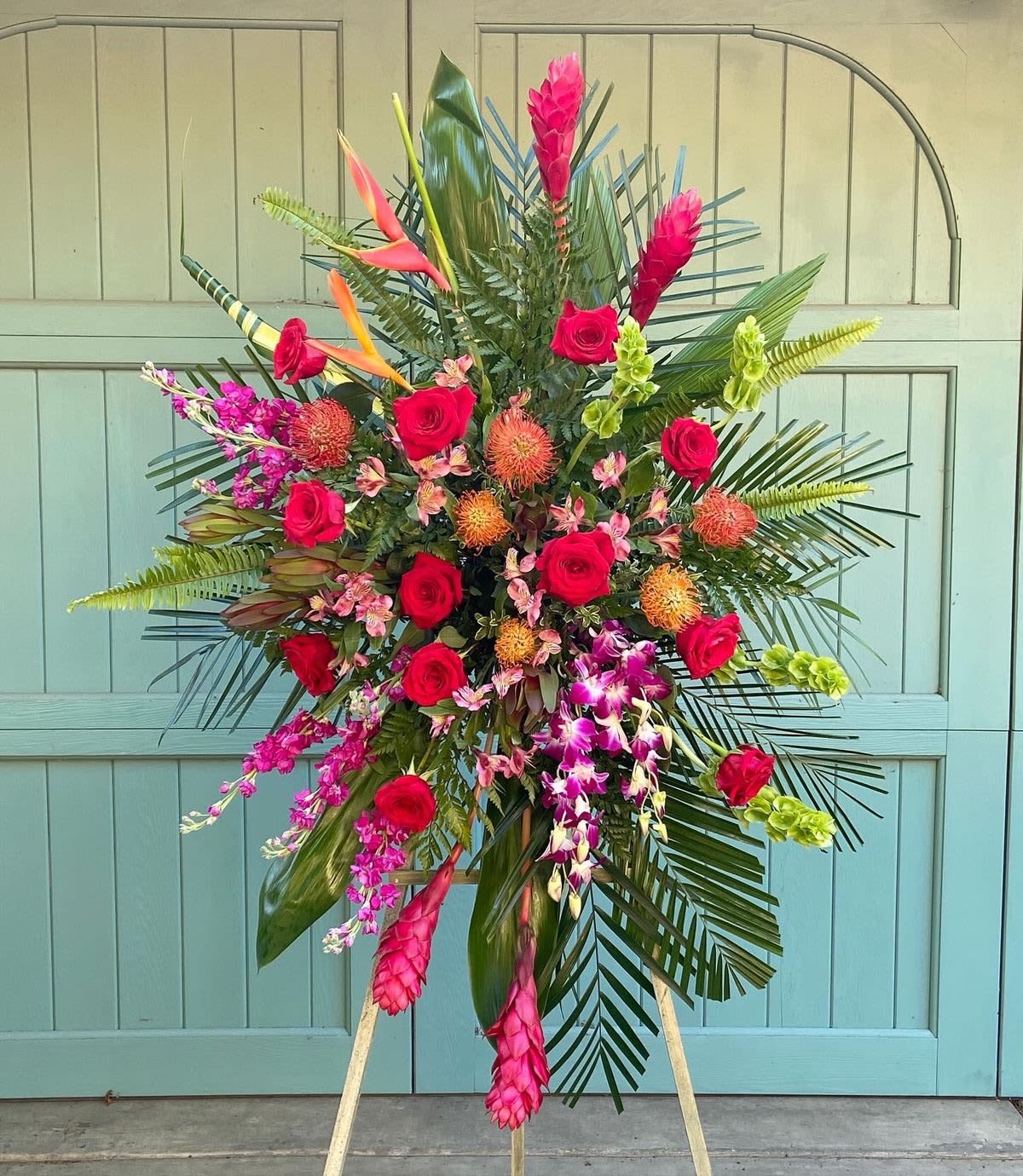 Tropical Tribute Standing Spray - A bold and vibrant expression of love and remembrance, the Tropical Tribute Standing Spray is designed to celebrate a life full of warmth, beauty, and spirit. This striking arrangement features an artful blend of lush tropical blooms and classic roses, creating a radiant display that feels both elegant and uplifting.  Rich red roses symbolize deep love and respect, while exotic pincushion protea, heliconia, and ginger add texture and a sense of natural beauty. Accents of cascading dendrobium orchids and delicate pink blooms soften the design, while dramatic palm fronds and tropical greenery create a full, fan-shaped silhouette.  Perfect for services, memorials, or celebrations of life, this standing spray offers a heartfelt tribute with a vibrant, island-inspired touch.