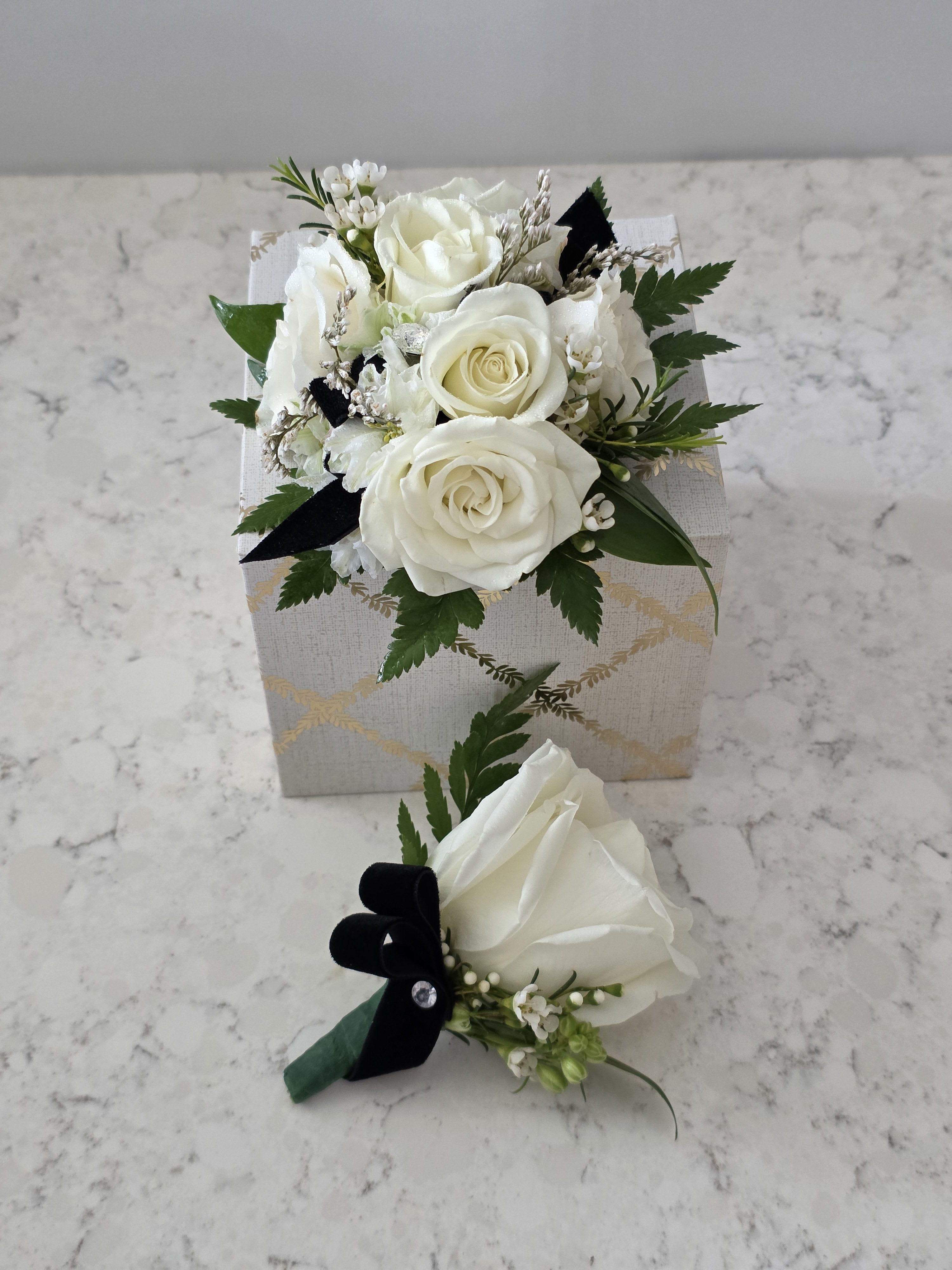 White Roses Corsage &amp; Boutonierre- Prom, Wedding, or Formals - A cluster of pure white roses tied with dark ribbon makes an arresting prom or wedding corsage and boutonniere set - the kind of small, elegant gift that makes the person you're honoring feel seen and celebrated. This formal arrangement works beautifully for proms, weddings, anniversaries, or any black‑tie event where crisp white blooms and tailored accents speak louder than words.  The compact corsage features several tightly grouped white roses with tiny white filler blossoms and fresh green sprigs, arranged on a patterned square gift box and finished with contrasting black ribbon for dramatic polish. The matching boutonniere is a single white rose with delicate filler and a black velvet ribbon bow, pinned securely with the included boutonniere pin. Styling hints in the images also show coordinating reception pieces - glass pedestal vases, yellow‑accent centerpieces, chair signs, and refined tableware - so this set pairs seamlessly with formal table décor.  Choose this set for a bride and groom, prom date, or honoree who prefers classic, understated florals; the restrained palette of white and green with bold ribbon contrast keeps the look timeless and easy to coordinate. Sizes are compact for wearability and presentation; each boutonniere includes a pin and the corsage is presented on a gift box for effortless gifting - order with same‑day options when available to ensure fresh delivery for your event.