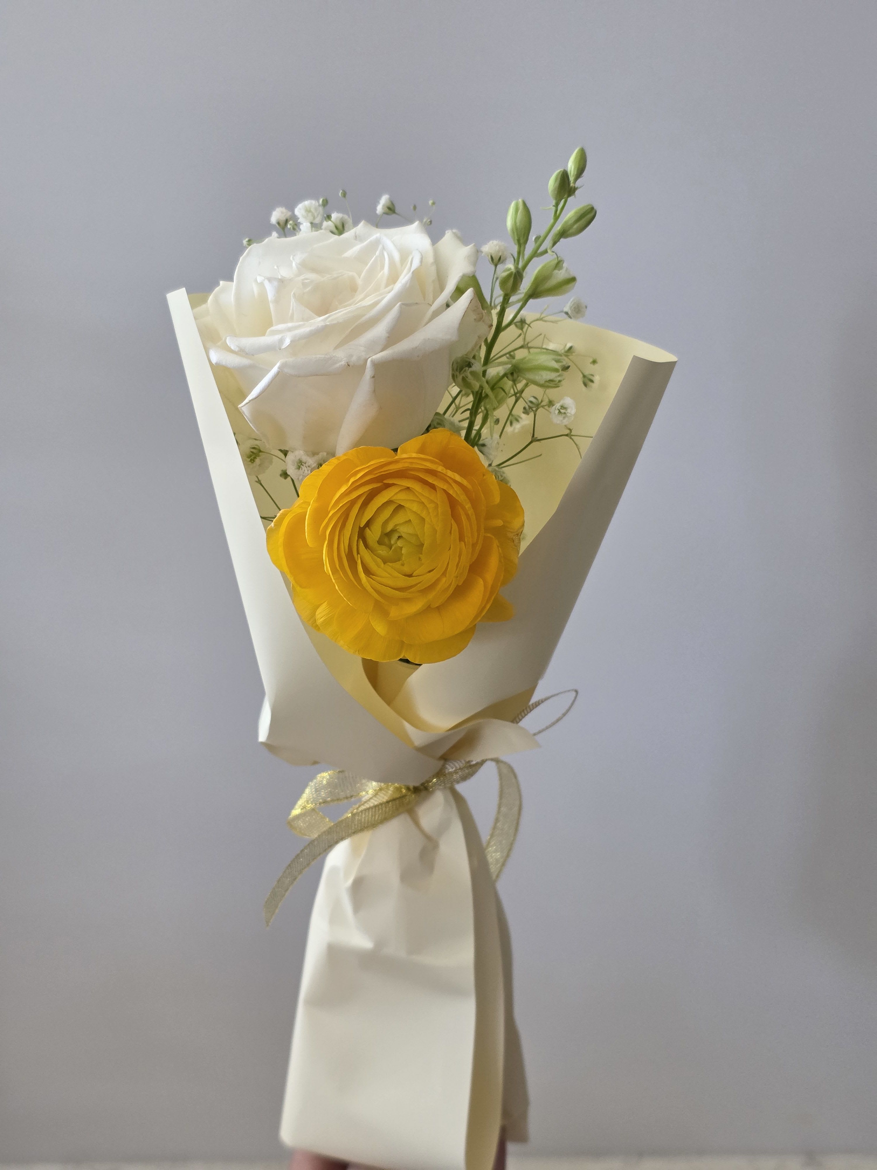 Teacher's Appreciation Day Bouquet  - A bright yellow ranunculus paired with a creamy white rose makes this Teacher's Appreciation Day bouquet a cheerful, "thank you" gift that feels both fresh and thoughtfully composed. Perfect as a thank-you or just-because surprise, the color mix-sunny yellow, clean white, and hints of red and blue from the playful apple ornament-reads lively without being fussy, so the recipient will feel warmly seen and appreciated.  Wrapped in soft cream paper and secured with a pale gold ribbon, the arrangement includes small white filler blooms and slender green buds for texture and movement. The small size is intentionally gift-ready and easy to carry to a classroom or desk; a glossy red glass apple ornament with a blue leaf adds a whimsical teacher-themed accent. Care is simple: keep stems in fresh water and out of direct midday sun to prolong vase life.