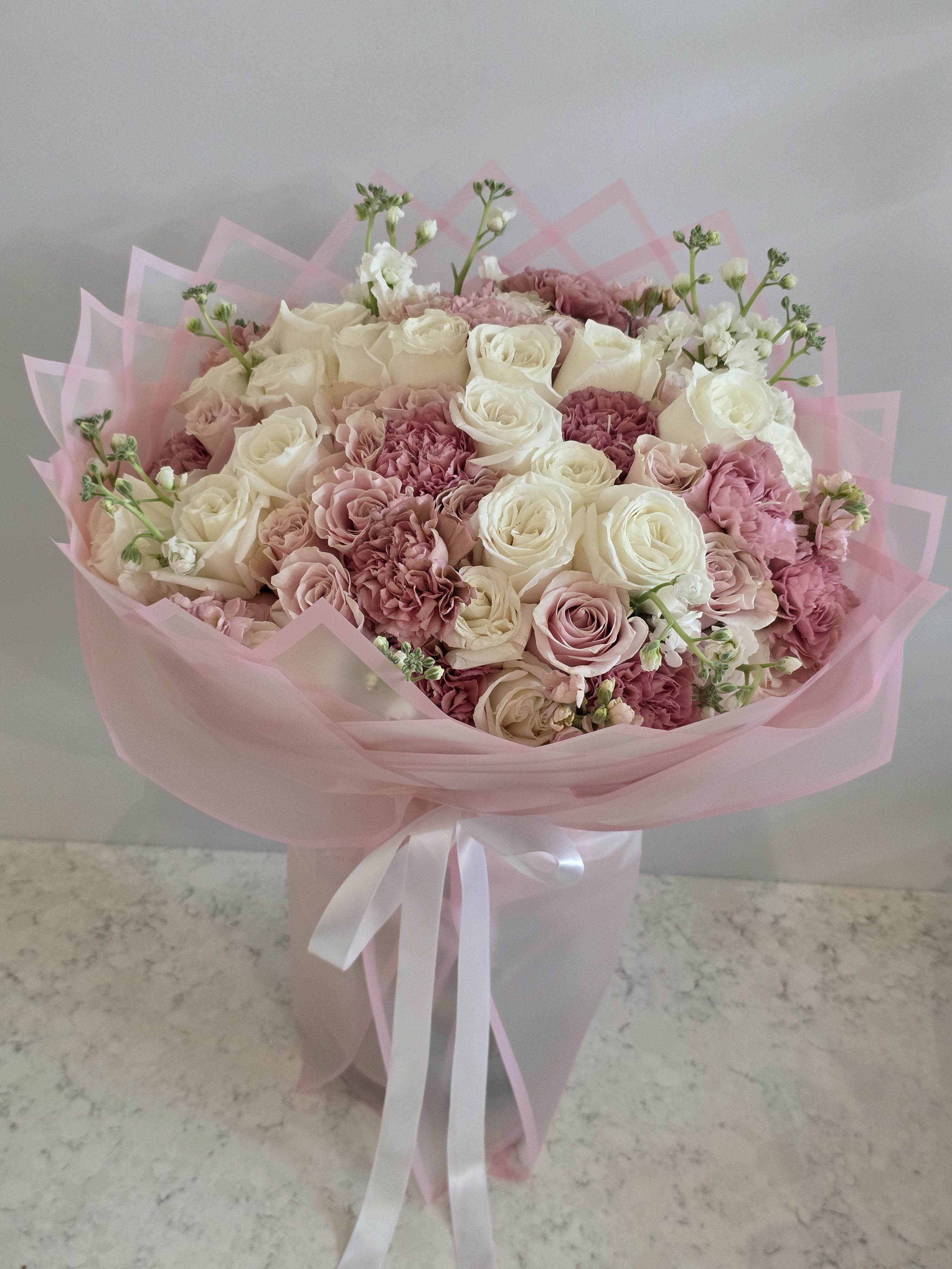 Pinky Promise Pink Rose Bouquet - 2 Dozen Roses - A lush, rounded bouquet of pink and white roses designed to make the person you're thinking of feel cherished - ideal as a birthday bouquet, anniversary flowers, or a Valentine's Day surprise. The Pinky Promise pink rose bouquet pairs two dozen roses with clusters of pink carnations and small pale blue filler blooms for a soft, romantic look that reads feminine and celebratory.  The arrangement is tightly gathered for a full, plush texture and wrapped in layered pink paper that adds a delicate, gift-ready finish. Whites brighten the deeper pinks while the pale blue accents add a whisper of contrast; the mix of roses, carnations, and filler creates both visual depth and gentle scent.  Perfect for saying "I love you," "happy birthday," or "congratulations," this bouquet arrives ready to present and will make the recipient feel truly seen and celebrated. For best display, trim stems and place in fresh water; order early for seasonal occasions to ensure availability.