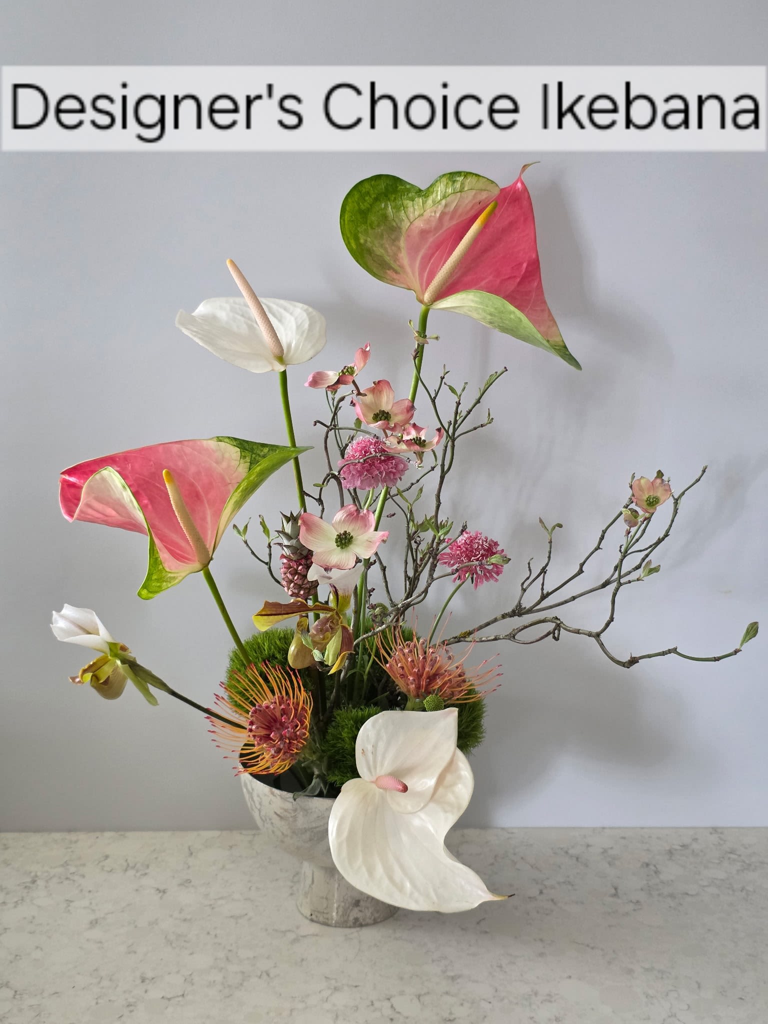 Designer's Choice Ikebana Arrangement  - A contemporary take on floral gift-giving, this Designer's Choice Ikebana arrangement brings artful balance to any room - perfect as a birthday bouquet, anniversary arrangement, or a thoughtful "thinking of you" gift. The design emphasizes sculptural lines and seasonal contrasts so the person you're sending it to feels noticed: one style is soft and serene for a calm, intimate impression, while the bolder version makes a vivid, exotic statement.  Arrangements feature mixed-color blooms with white and pink branching blossoms in a low ceramic bowl set over moss and decorative branches for an airy, delicate silhouette, or bright orange tropical flowers and orchids paired with purple accents and broad, fan-like leaves in a dark, black vase for a striking, architectural look. Each composition uses quality hard goods - ceramic bowls or black vases, moss, and accent branches - to hold the floral form and create lasting visual impact.  Choose the softer design to soothe and celebrate quiet moments, or the vivid tropical version to surprise and energize someone who loves bold color and texture. Sizes and exact flower varieties will reflect seasonal availability as these are designer's choice arrangements crafted to highlight freshness and composition.