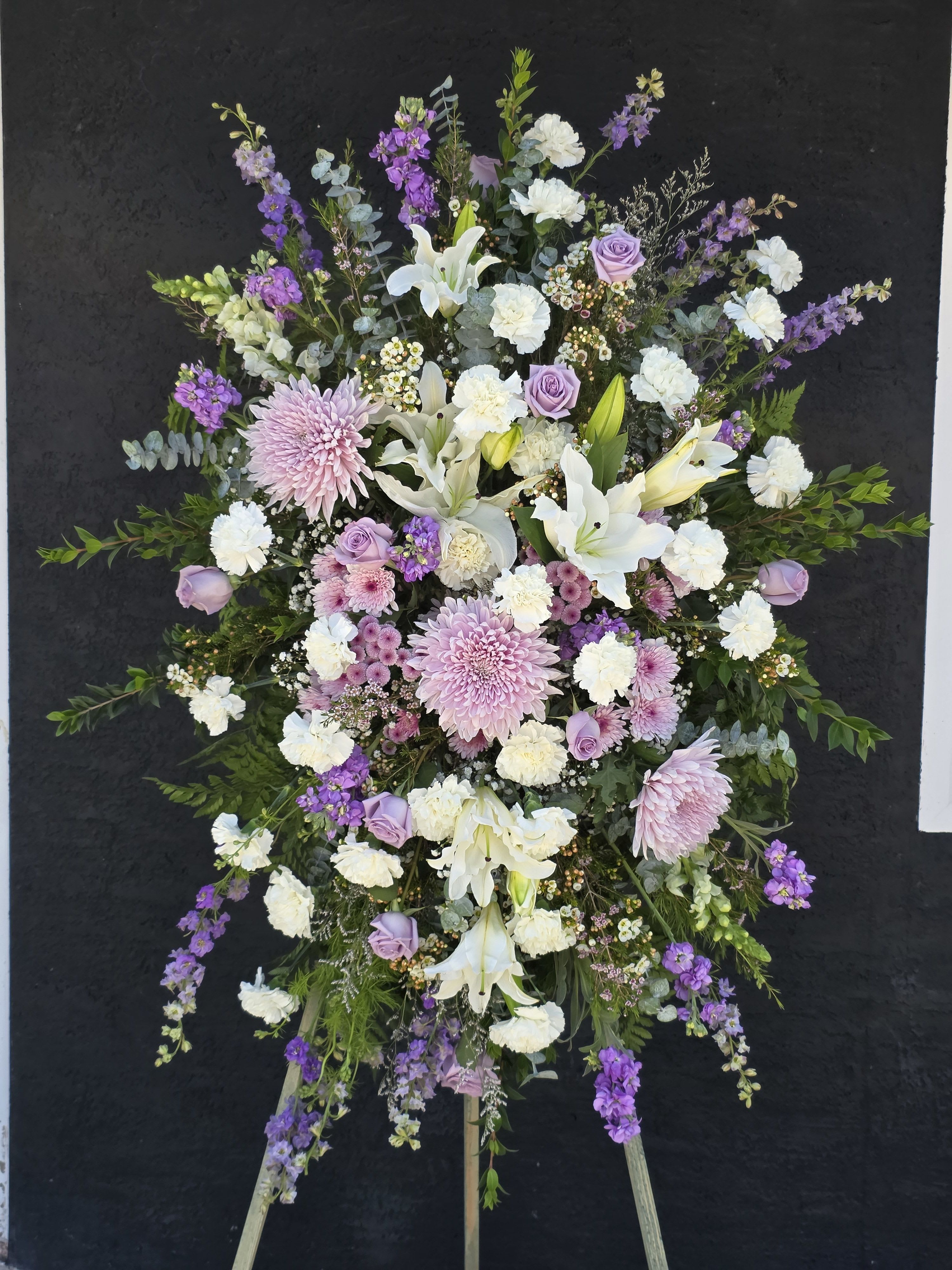 Lilac Funeral Standing Spray - Dense pink lilies and clusters of red and blush roses create the first impression-a lush standing spray designed for sympathy and funeral services that reads as both romantic and respectfully traditional. This large oval arrangement pairs white chrysanthemums and pale lavender filler blooms with broad palm fronds for a formal, ceremonial silhouette; a white ribbon bow and printed condolence streamers at the front complete the memorial presentation so the person you're honoring will feel truly remembered.  Built on a sturdy white easel and standing-spray frame, the tribute is full and layered with carnations, spray roses, stock, and other seasonal accents tucked among the primary blooms for texture and depth. The palette of pink, white, red and soft purple yields a comforting contrast that photographs well and carries across indoor service spaces or graveside displays. Orders arrive ready to display on the included easel; please add special wording for the printed streamers or delivery instructions at checkout so we can prepare the tribute exactly as you intend.