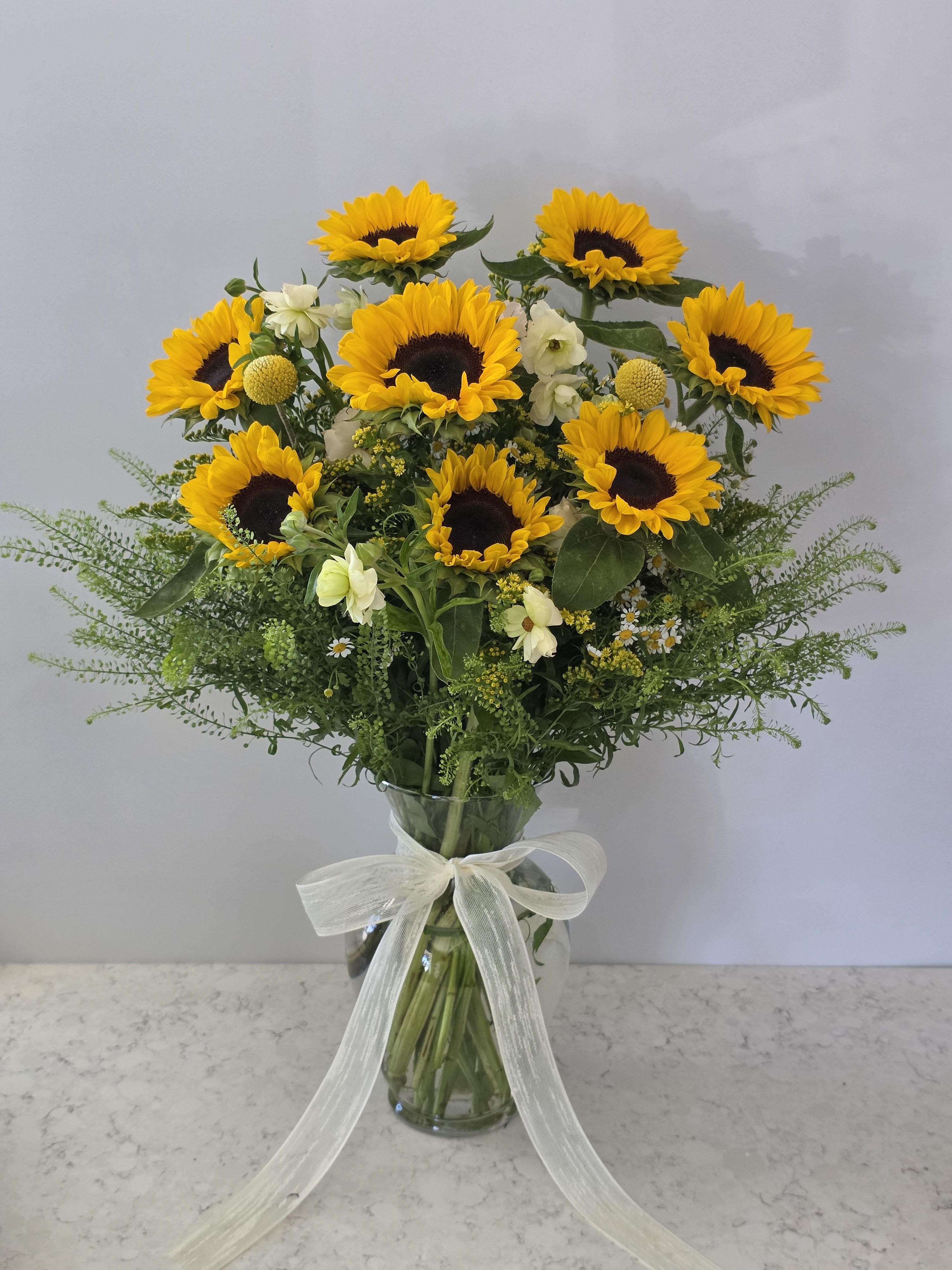 Stunning Sunflowers Arrangement  - Golden sunflowers steal the show in this vibrant hand-tied flower arrangement, a joyful birthday bouquet and anniversary gift that also reads perfectly for "just because," thank-you, or romantic moments. Hot pink and blush roses mingle with vivid purple irises and small white daisies for a bold, celebratory palette-warm yellows, cool violets, and soft pinks that make the recipient feel genuinely seen and celebrated from the moment it arrives.  Tightly clustered into a rounded, abundant shape, the blooms sit either as a wrapped bouquet with a decorative handle finished in lace and burlap or arranged upright in a clear glass vase tied with a sheer white ribbon-both presentations are gift-ready. Expect lush roses, sunny sunflowers, slender irises, airy greenery, and white filler blooms for texture; the mix reads lively and garden-fresh without being overly formal.  Perfect for spring weddings, Mother's Day, congratulations, or a romantic surprise, this arrangement is sized to feel generous while fitting easily into most vases. To keep blooms at their best, place in fresh water and recut stems every few days-an effortless, uplifting way to mark a milestone or brighten an ordinary day.