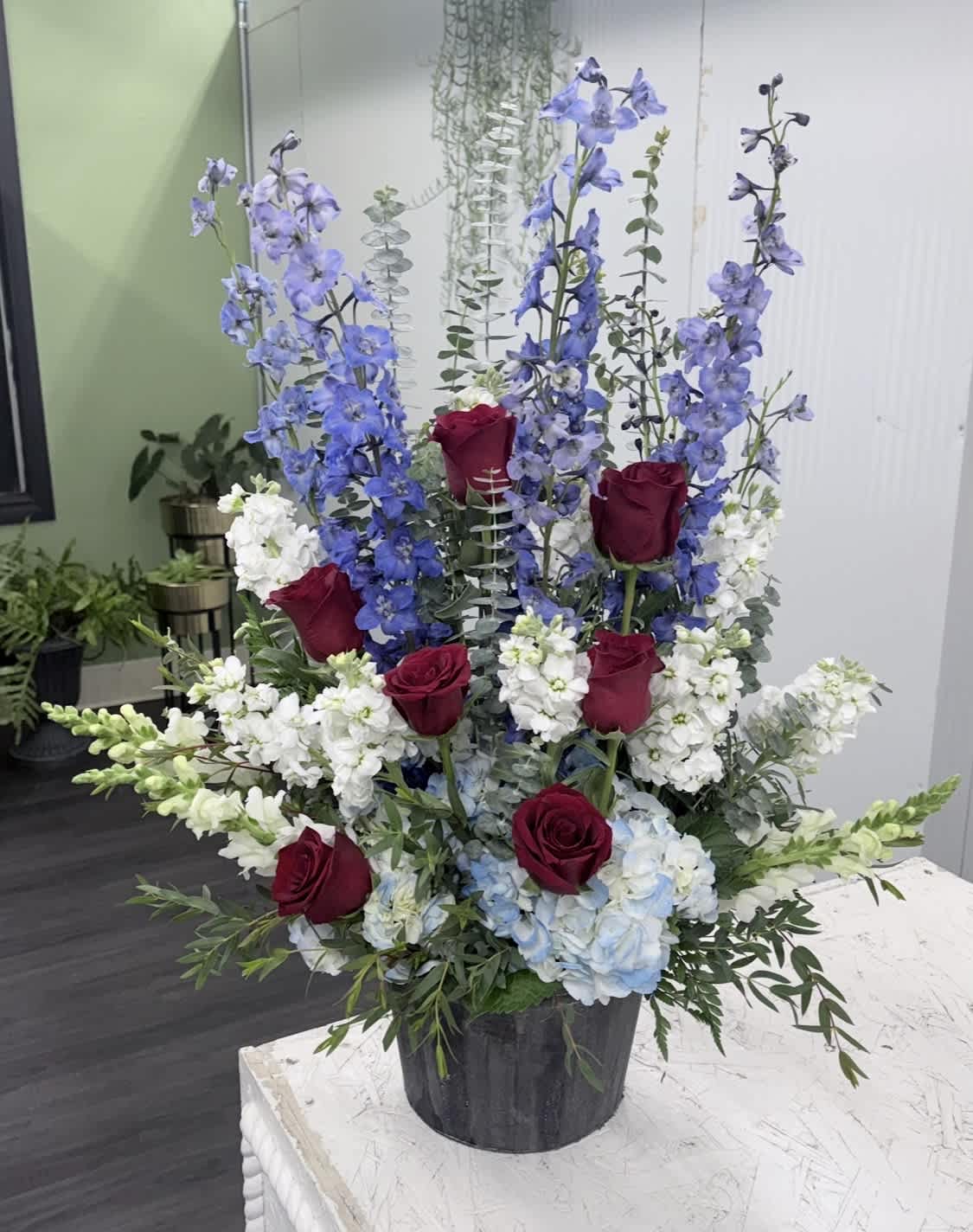 Tall floral arrangement with red roses, blue delphinium, and white blooms in a black container