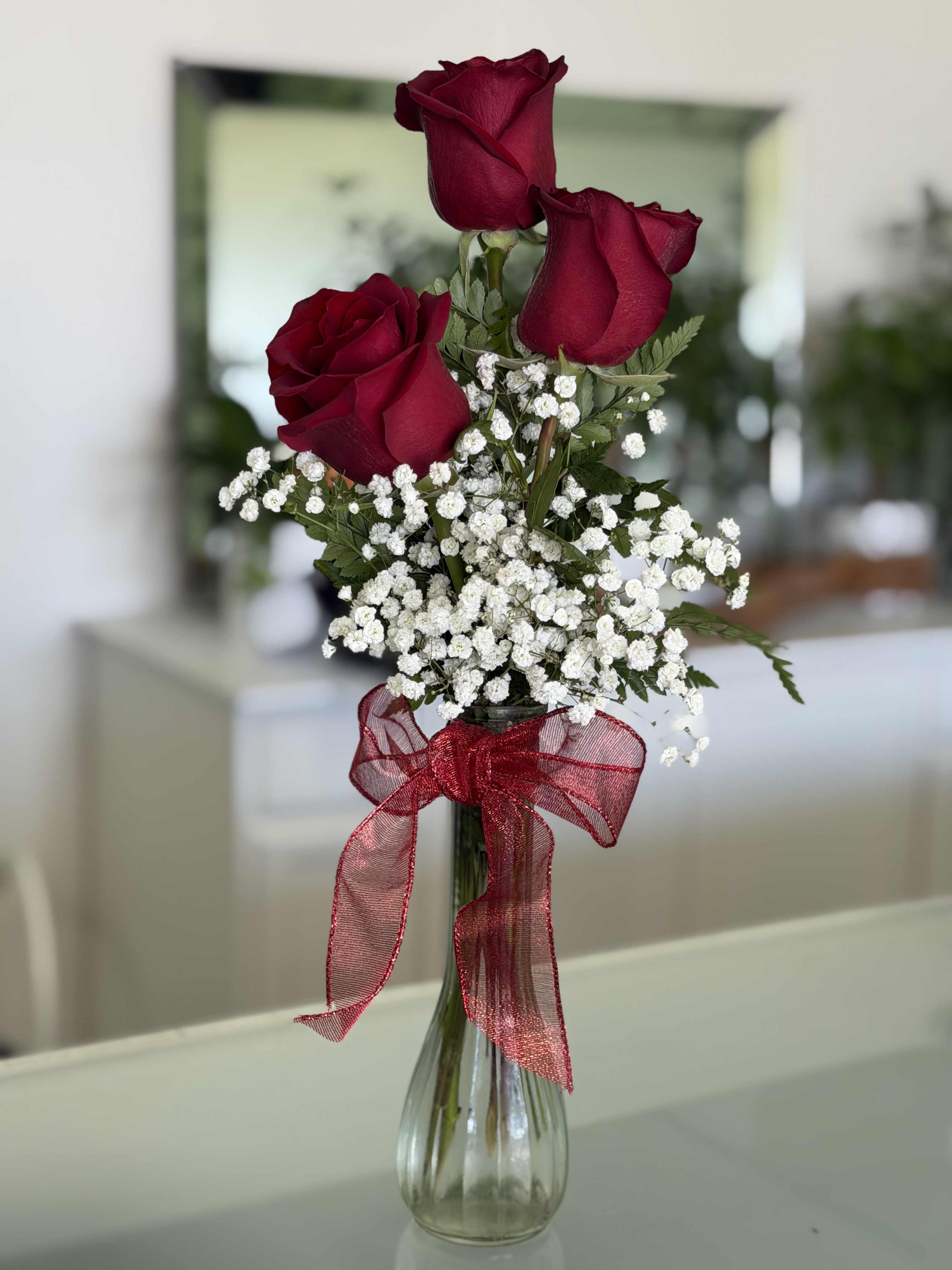 Three red roses in a glass vase with white baby's breath and a red ribbon